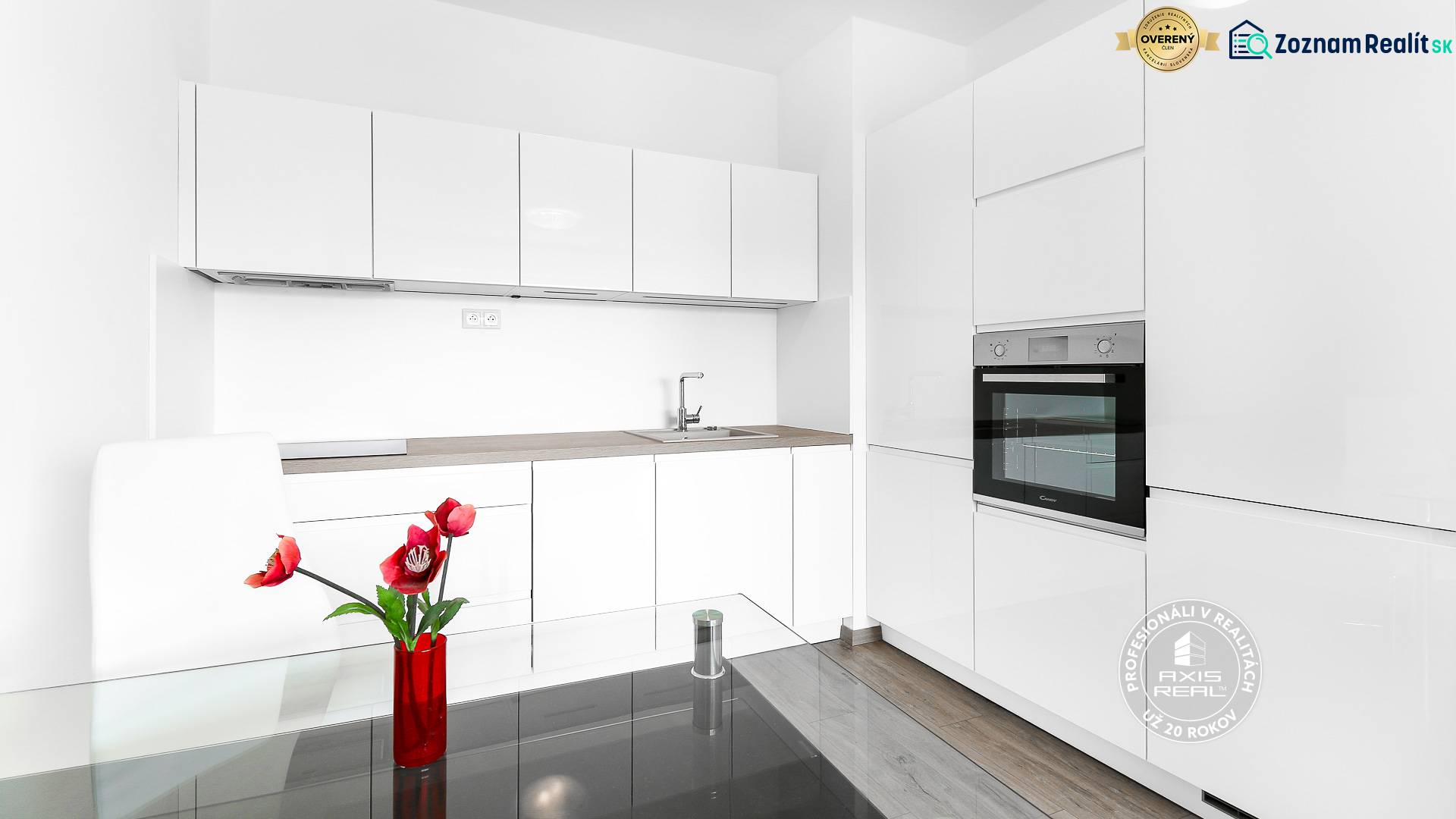 Kuchyně s bílými skříňkami v dvoupokojovém bytě, podlaha s dřevěným dekorem, červené květiny.