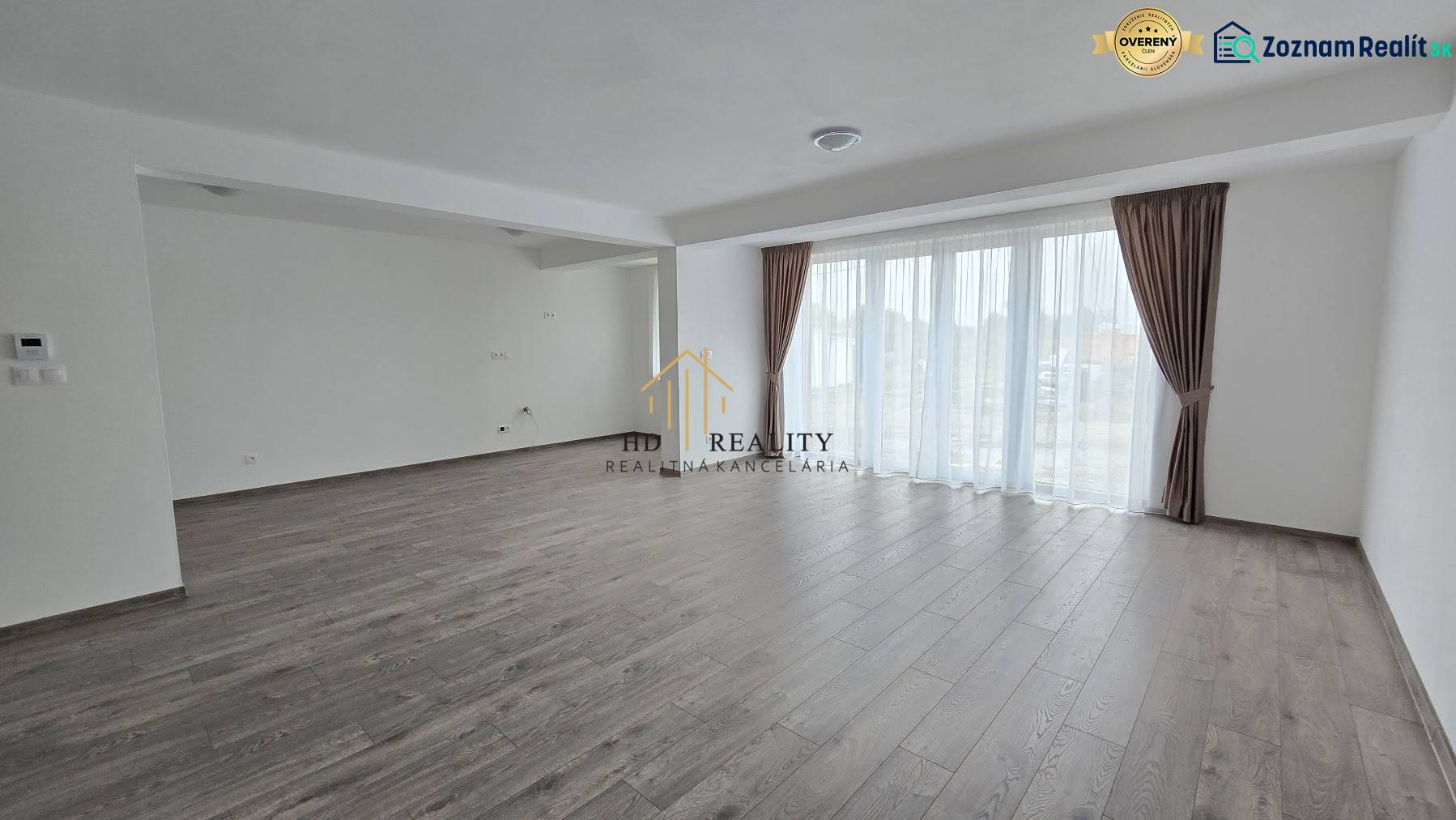 Obývací pokoj v rodinném domě s podlahou a světlými stěnami a velkými okny.