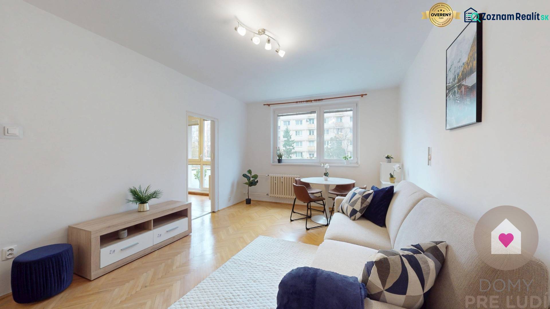 Obývací pokoj 1+kk bytu s podlahou s dřevěným dekorem, gaučem a kulatým stolem.