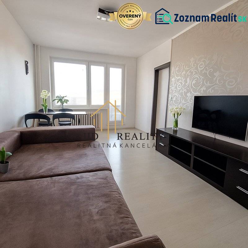 Obývací pokoj v 3-pokojovém bytě s velkou sedačkou, TV a podlahou s dřevěným dekorem.