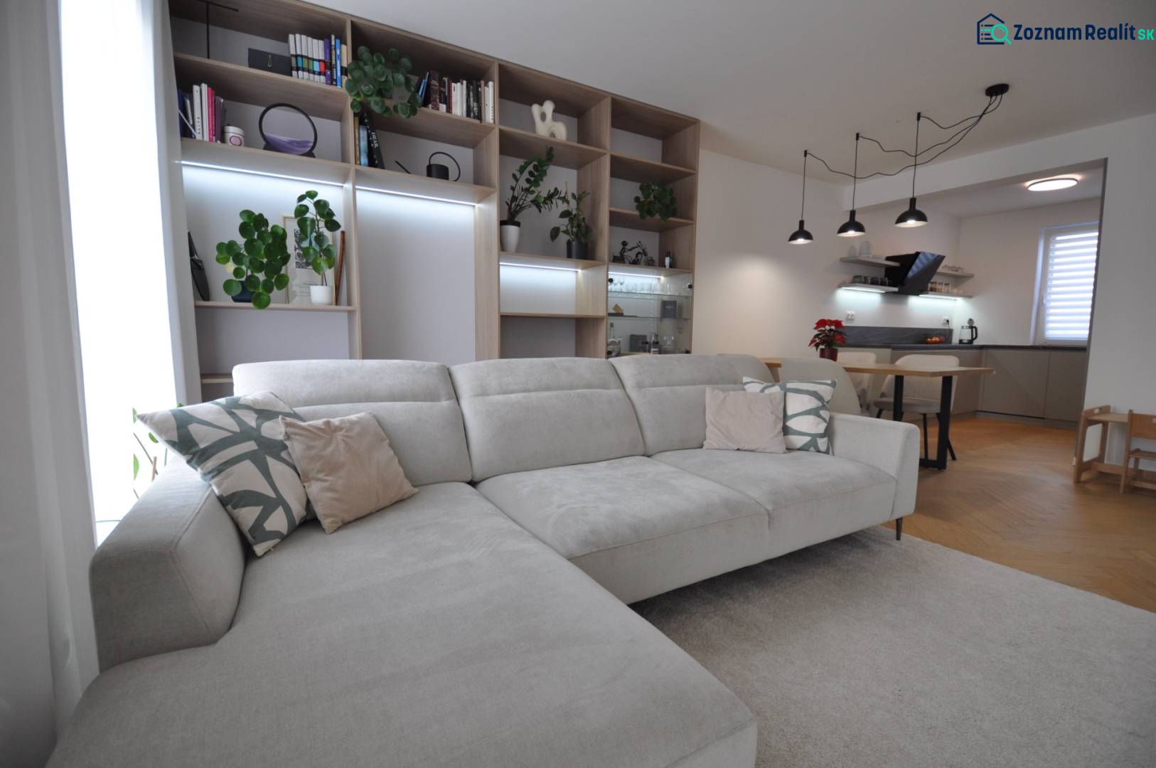 Obývací pokoj s velkou pohovkou, knihovnou a kuchyňským koutem v rodinném domě.