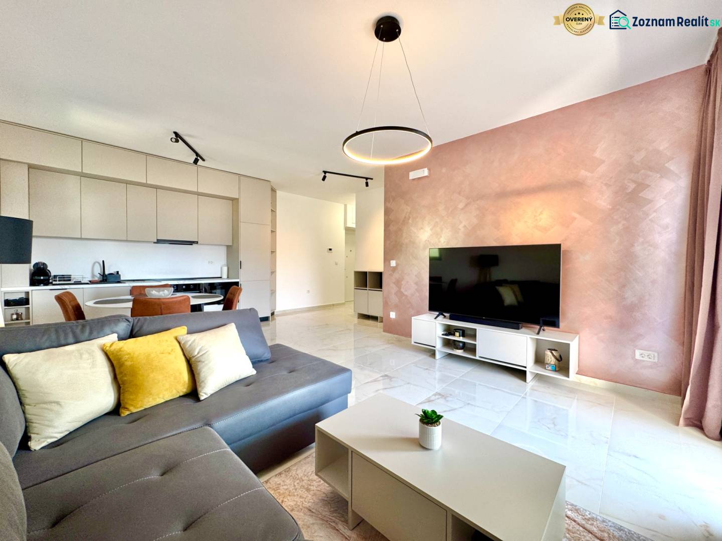 Obývací pokoj v 1+kk bytě s kuchyní, šedou sedačkou, televizorem a jídelním prostorem.