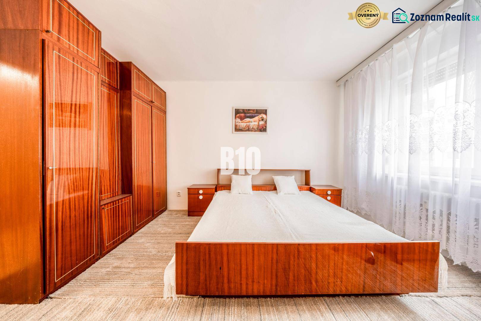 Ložnice ve 4-pokojovém bytě s manželskou postelí a vestavěnými skříněmi, světlé záclony.