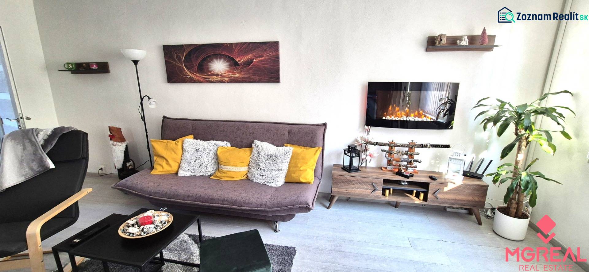 Obývací pokoj v 2-pokojovém bytě s pohovkou, policí a elektrickým krbem.