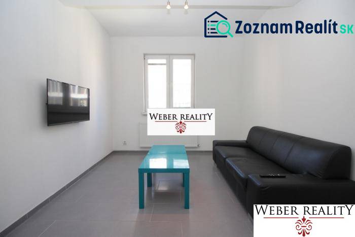 Obývací pokoj dvoupokojového bytu s černou sedačkou, televizorem na stěně a nízkým stolkem.