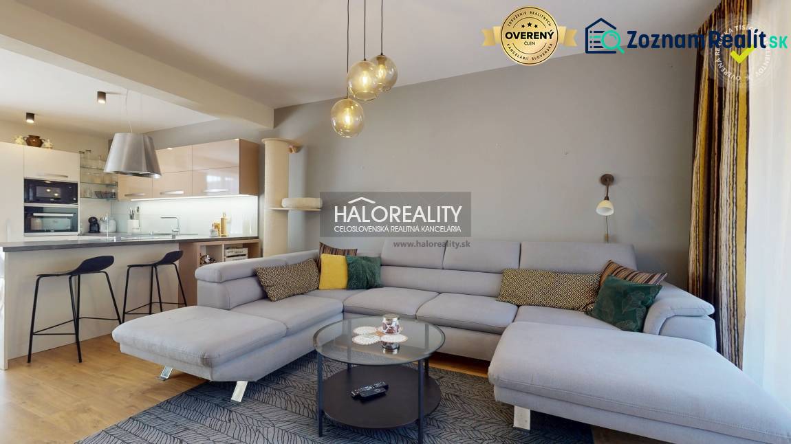 Prostorný obývací pokoj v rodinném domě s pohovkou, kuchyňským koutem a podlahou s dřevěným dekorem.