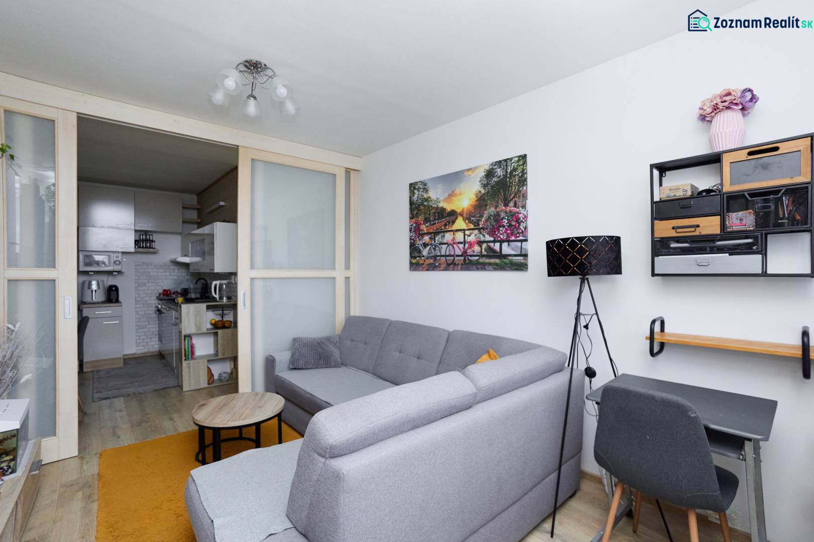 Obývací pokoj v 2-pokojovém bytě s podlahou s dřevěným dekorem, šedou pohovkou a přístupem do kuchyně.