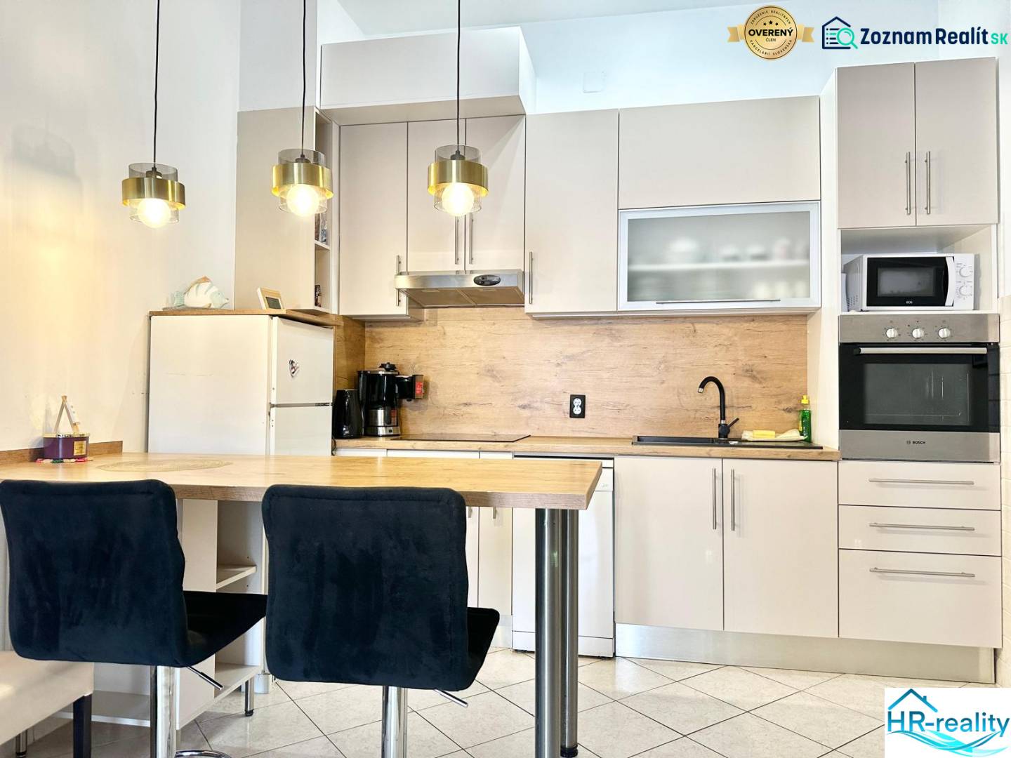 Kuchyně v rekreačním apartmánu s barovým pultem, závěsnými svítidly a bílou skříní.