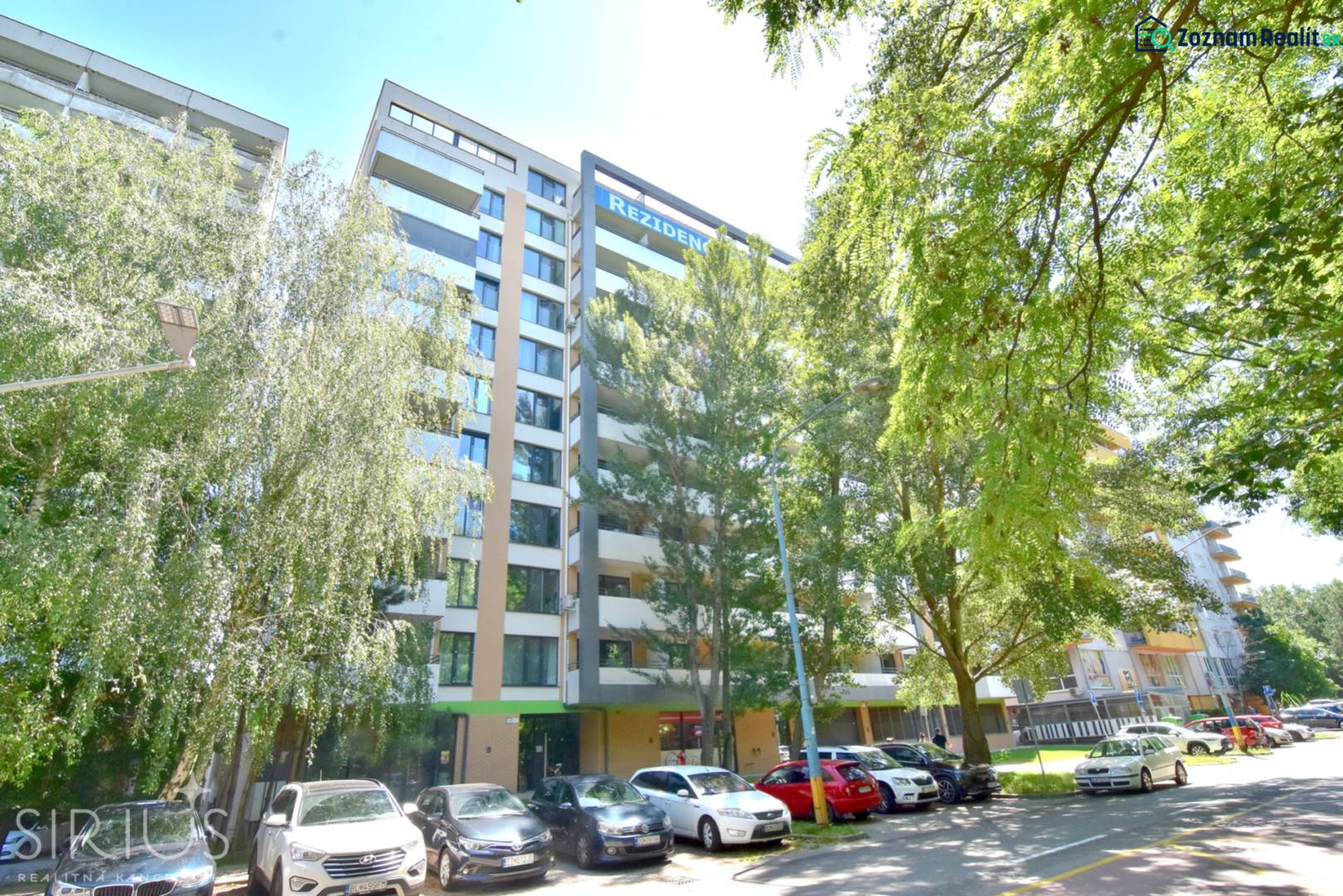 Bytový dům na Antolské ulici v Bratislavě - Petržalce, obklopený zelení a parkujícími auty.