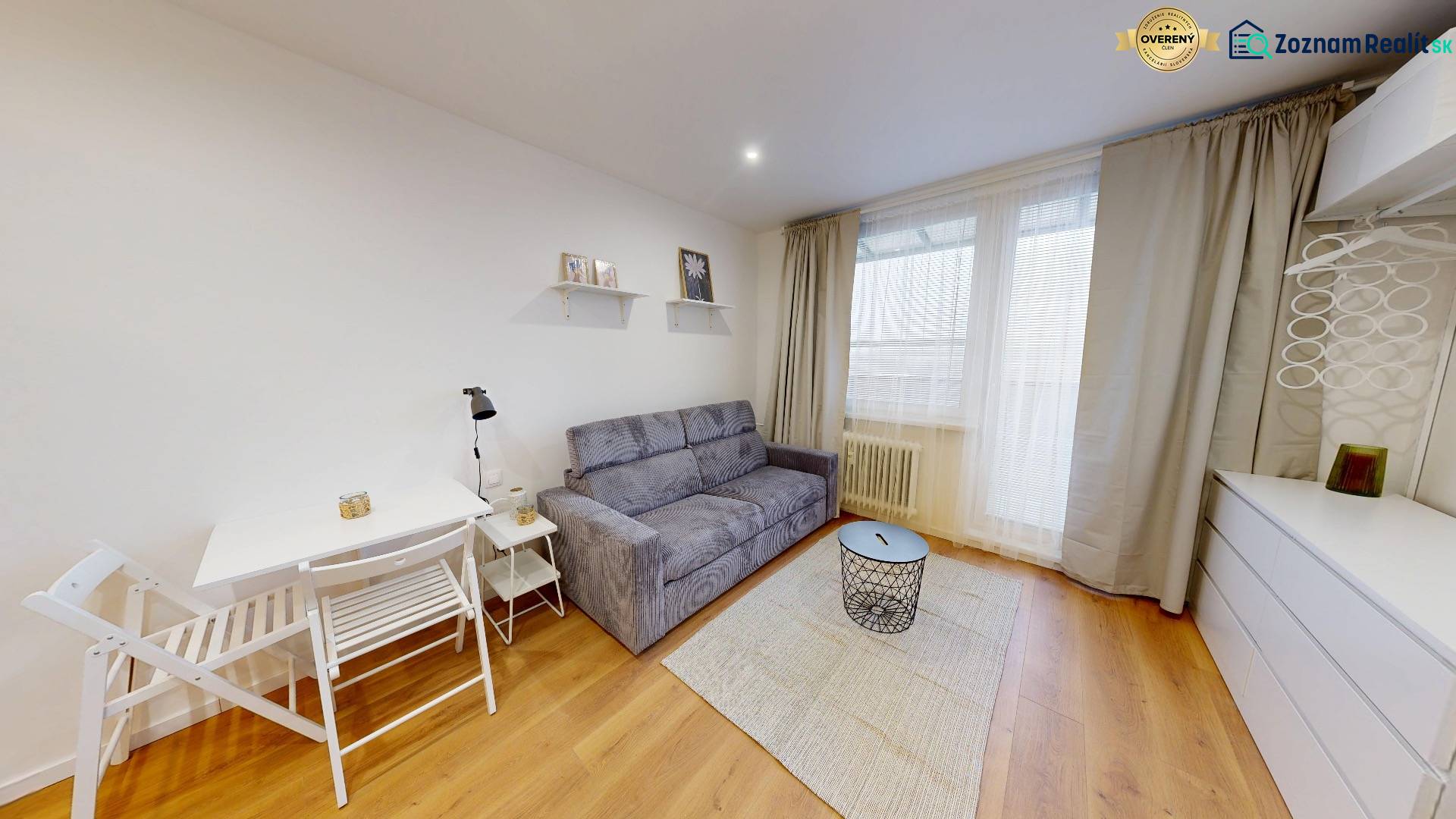 Garsonka s podlahou s dřevěným dekorem, sedačkou, stolkem a bílým nábytkem.