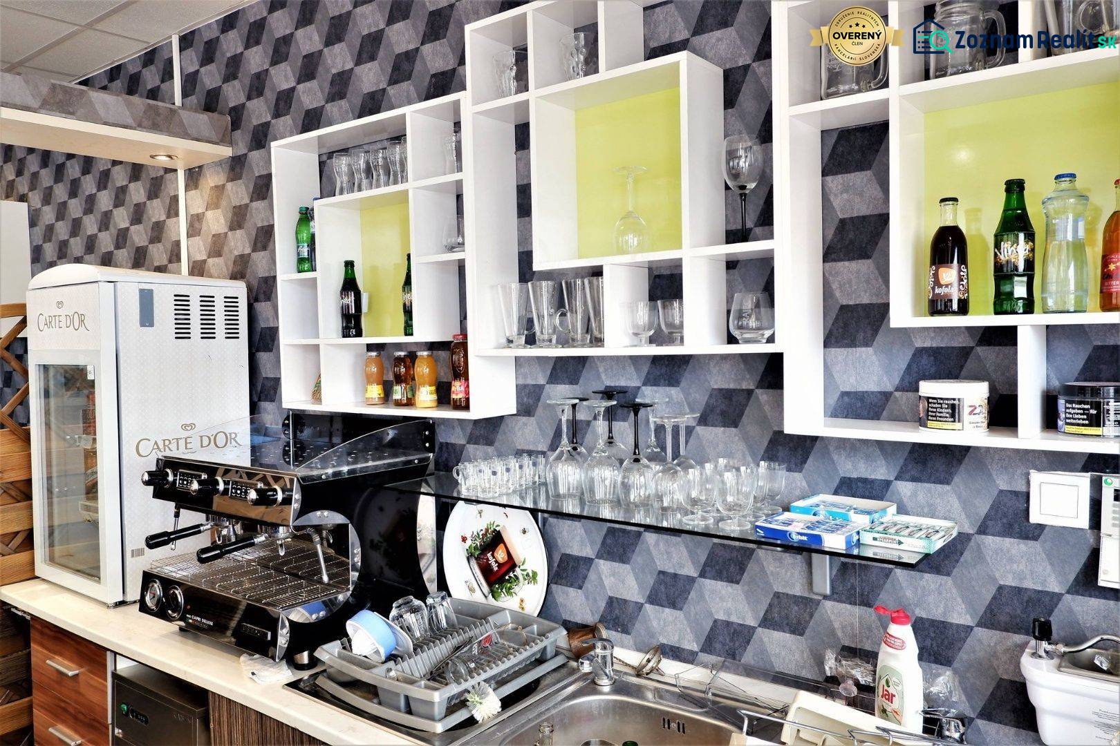 Interiér obchodních prostor s kávovarem, regály, sklenicemi a nápoji na poličkách.