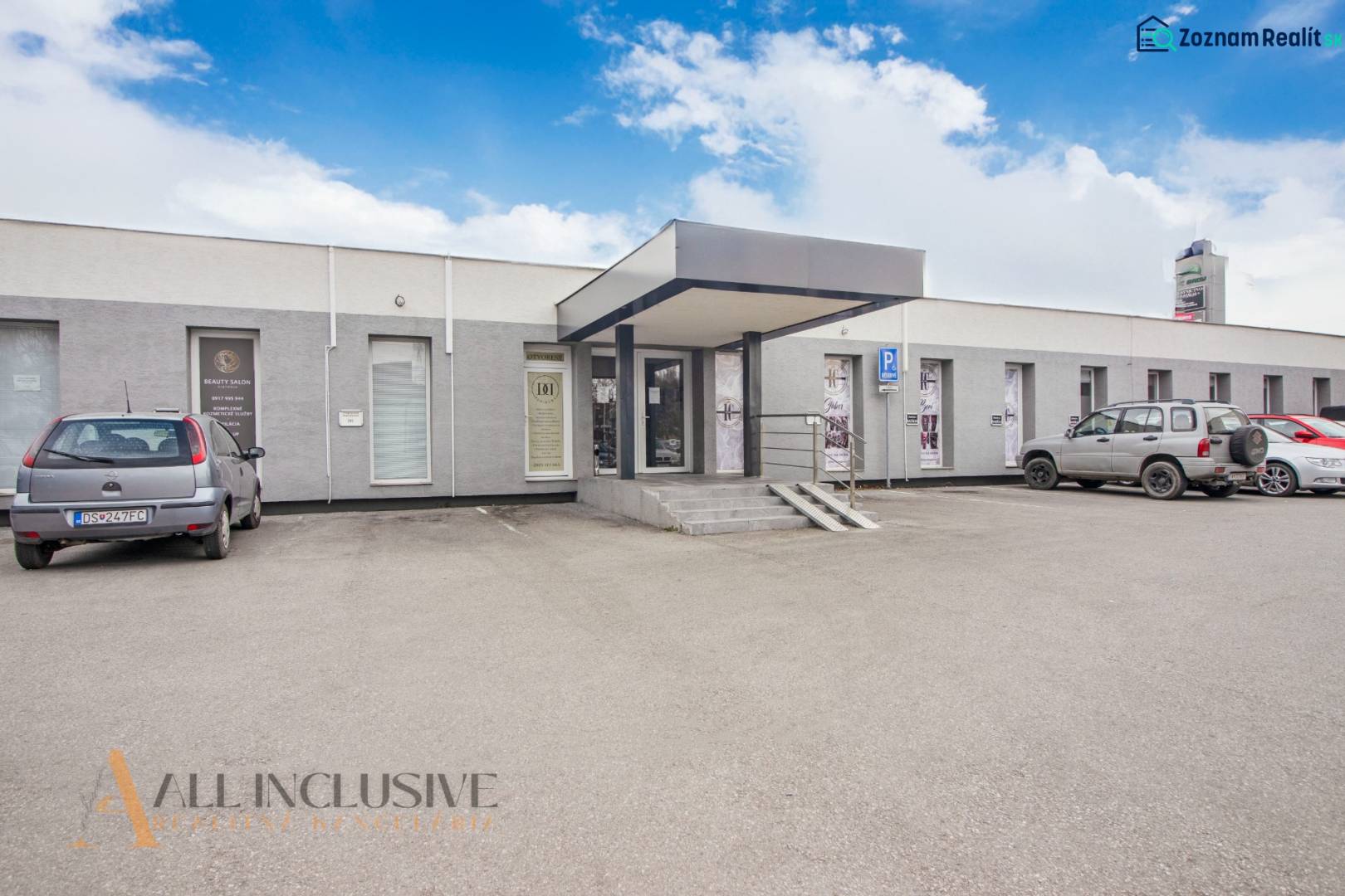 Jednopodlažní budova s obchodními prostory na ulici Vajanského, parkovištěm a auty před vstupem.