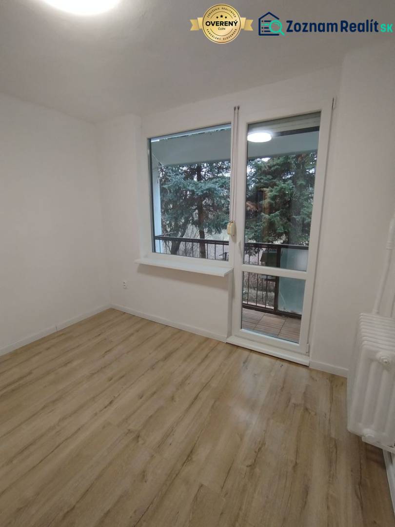 Pokoj se světlými stěnami, podlahou s dřevěným dekorem a výhledem na zeleň.