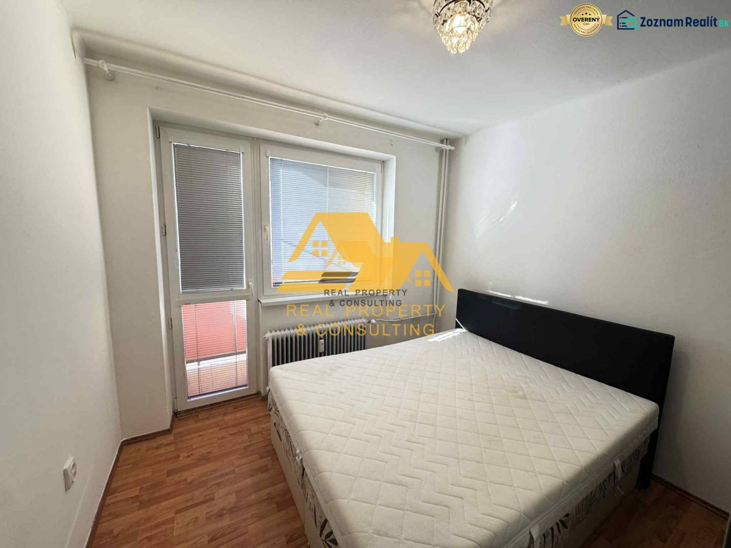 Ložnice ve 3pokojovém bytě s postelí, oknem a podlahou s dřevěným dekorem.