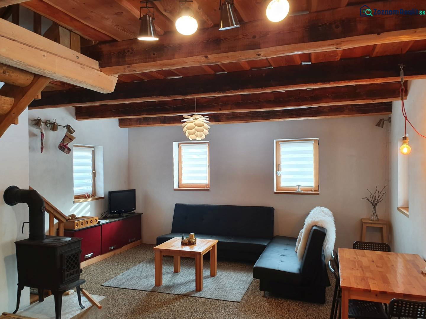 Interiér chaty s kachlovými kamny, pohovkou a dřevěným stropem z masivních trámů.