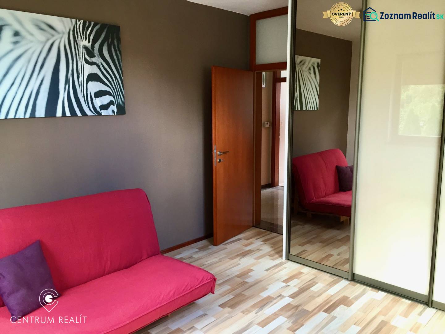 Pokoj s růžovou pohovkou, obraz zebry a zrcadlová skříň, podlaha s dřevěným dekorem.