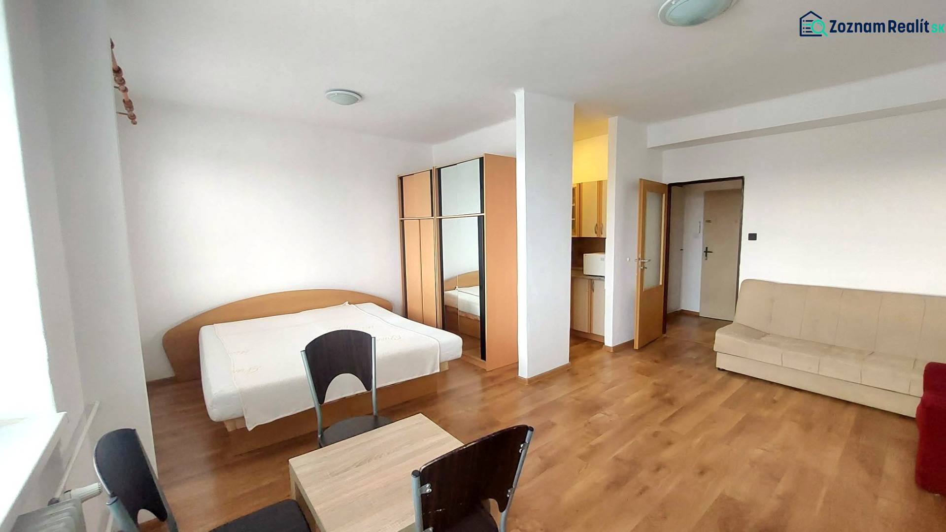 1+kk byt s podlahou s dřevěným dekorem, postelí, gaučem a skříní.
