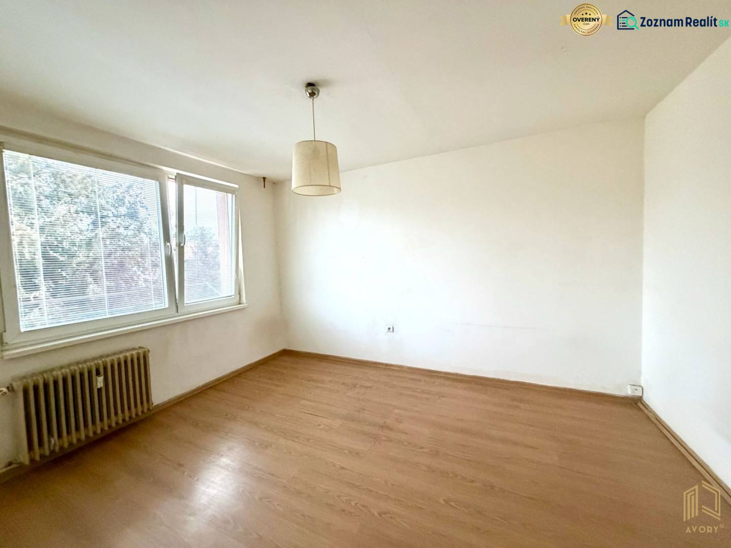 1+kk byt s velkým oknem, podlaha s dřevěným dekorem, bílá stěna, radiátor.