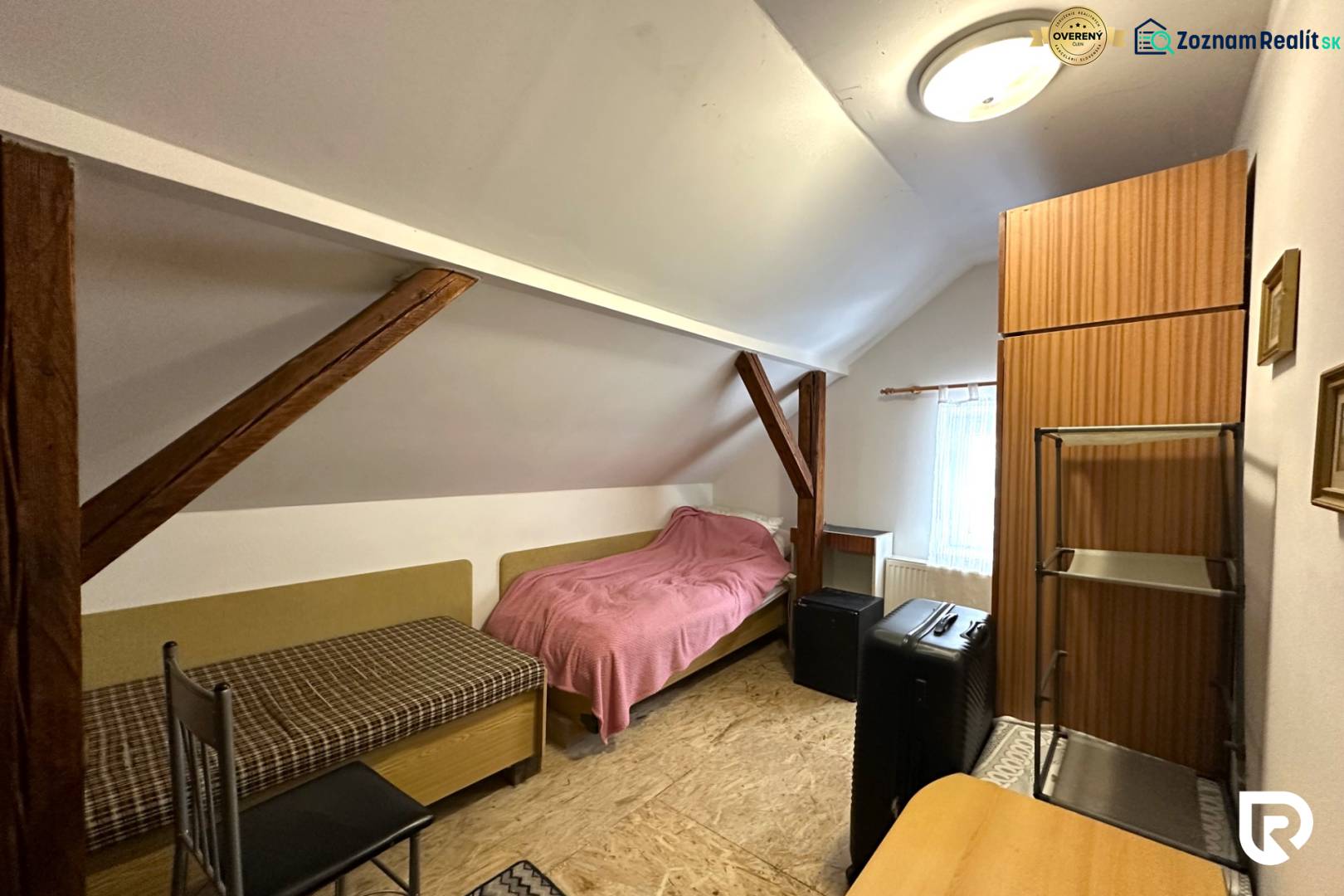 Podkrovní pokoj v rodinném domě s jednoduchým nábytkem a střešními trámy.