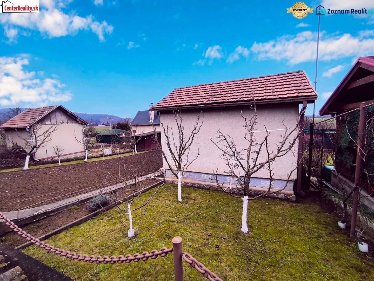 Zahrada s chatou v Partizánském, ovocné stromy a obdělávaná půda, jasná obloha.