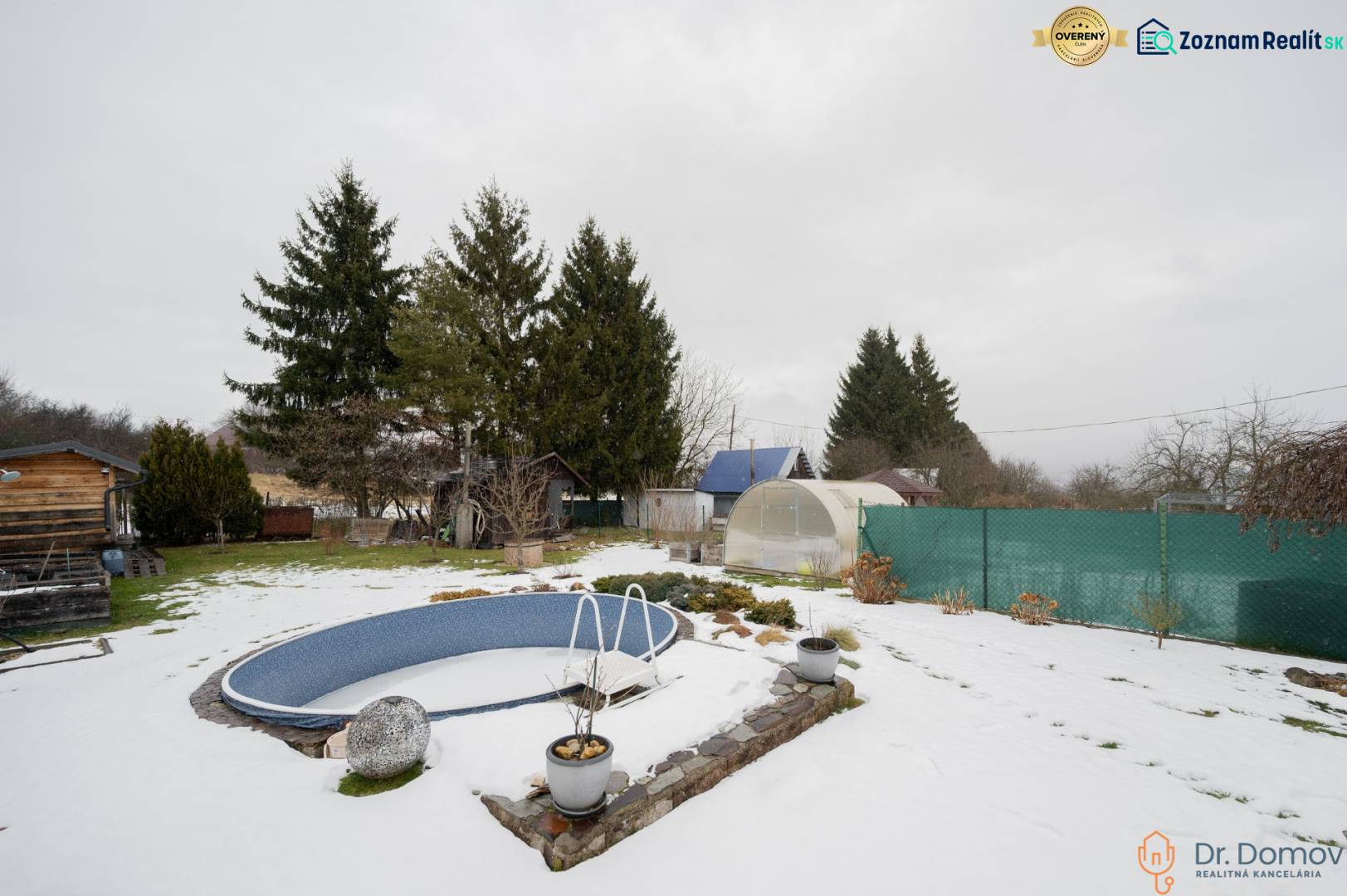 Zasněžená zahrada u chaty v Handlové s bazénem, skleníky a jehličnatými stromy.
