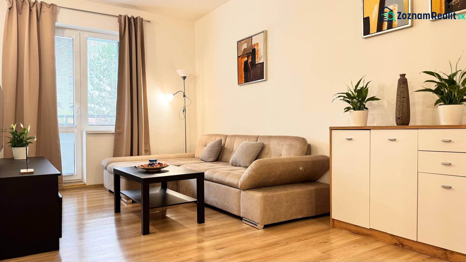 Obývací pokoj v 2pokojovém bytě s béžovou sedačkou, podlaha s dřevěným dekorem.