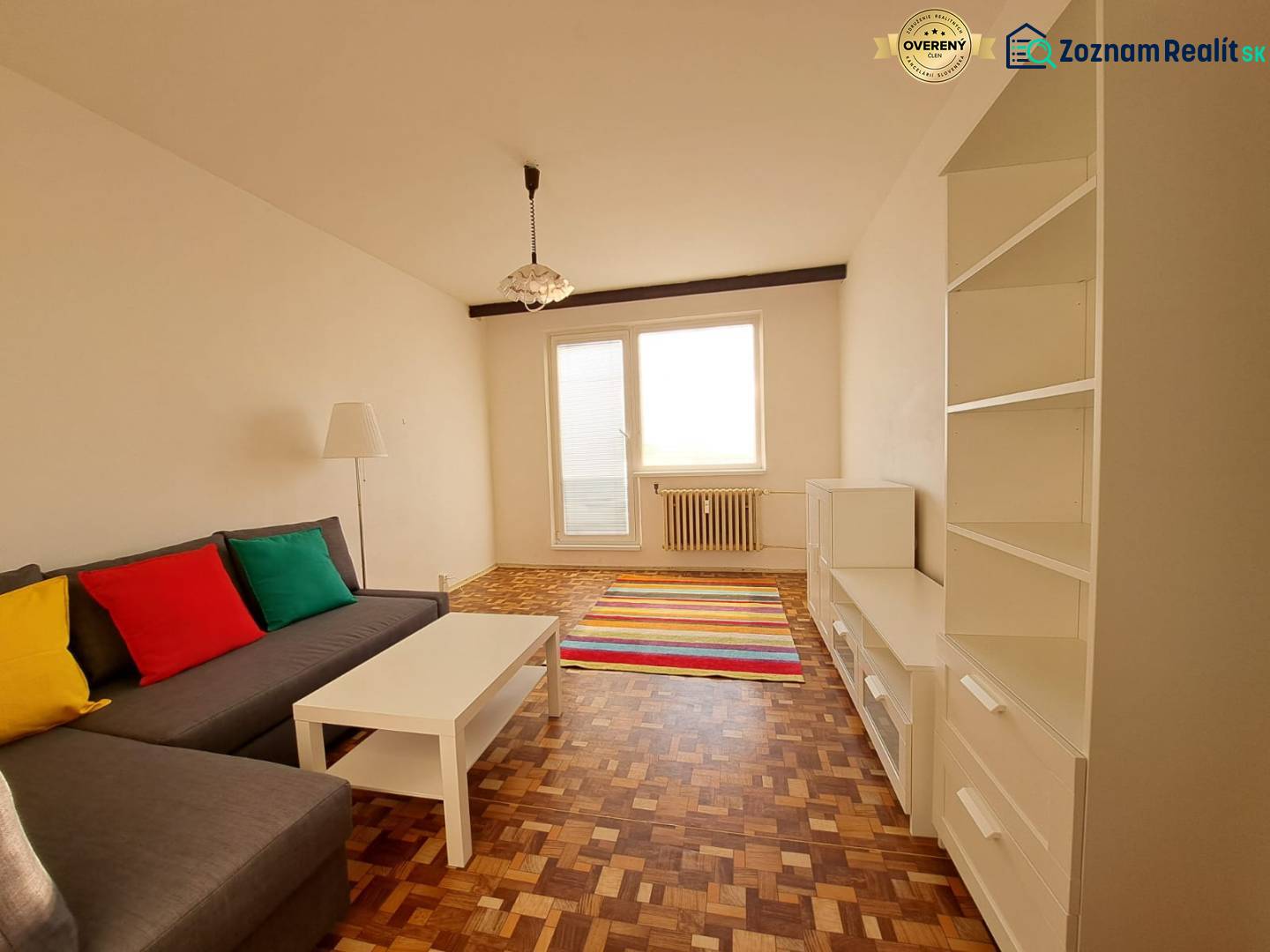 Obývací pokoj v 2-pokojovém bytě s pohovkou, bílým nábytkem, pestrobarevnými polštáři a podlahou s dřevěným dekorem.