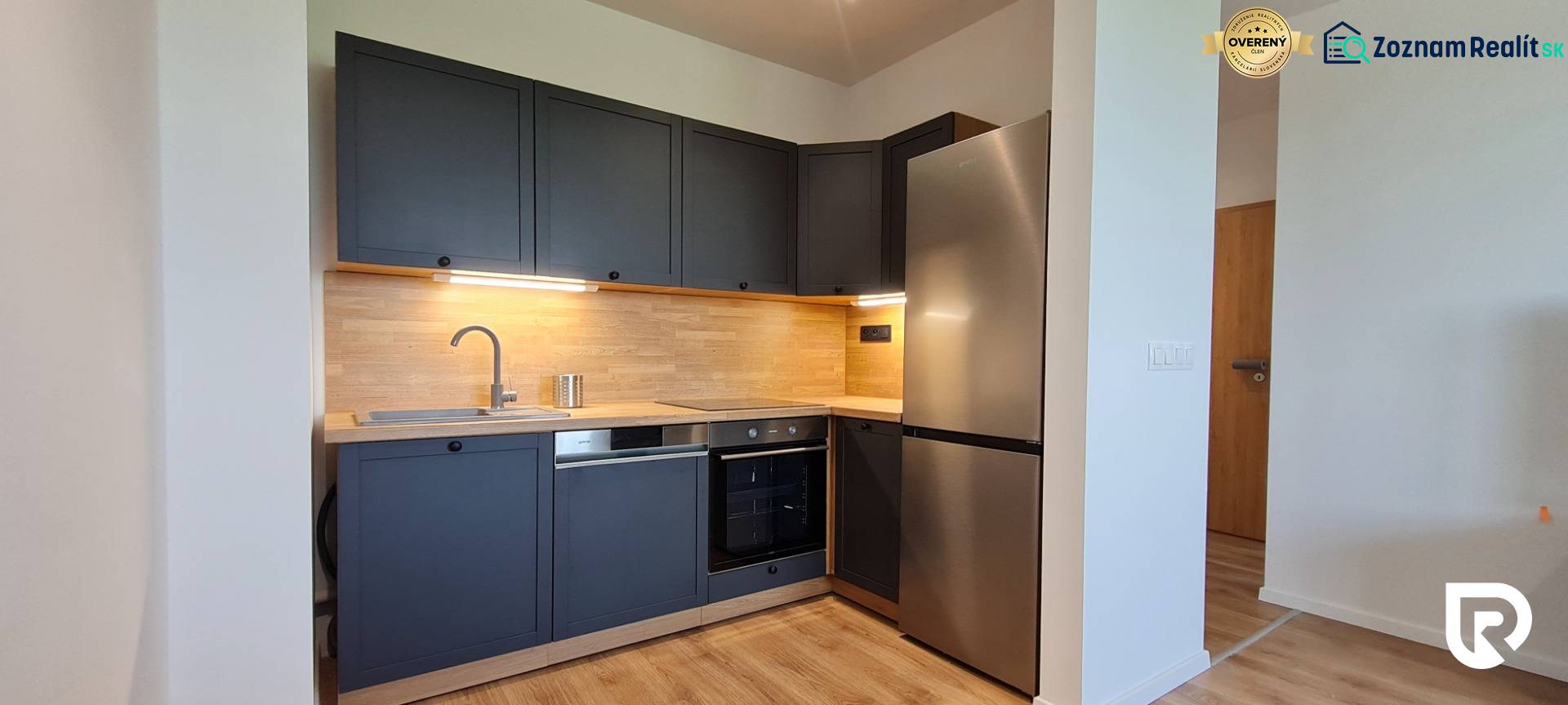 Kuchyně v 2pokojovém bytě s tmavě šedými skříňkami, dřevěným dekorem a nerezovými spotřebiči.
