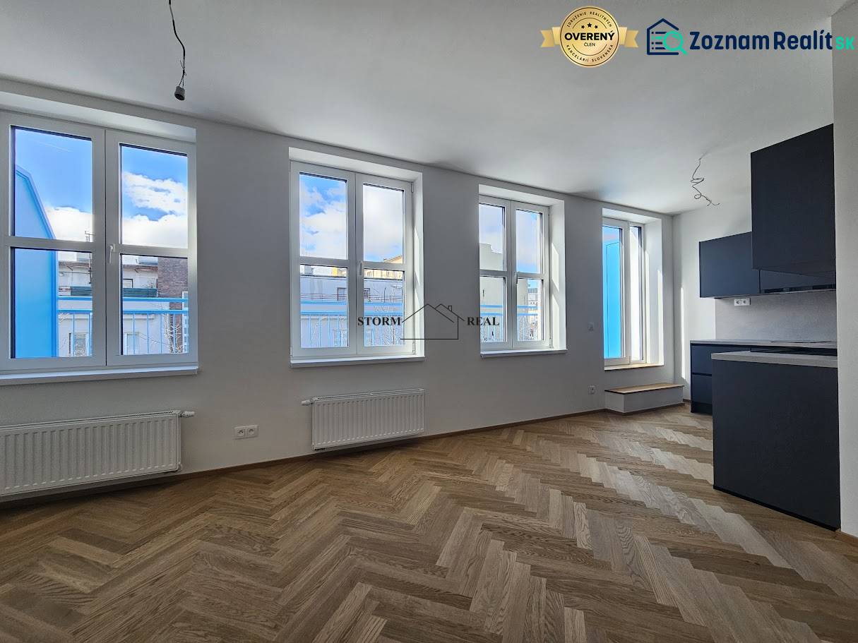 1+kk byt s podlahou s dřevěným dekorem a černou kuchyňskou linkou.