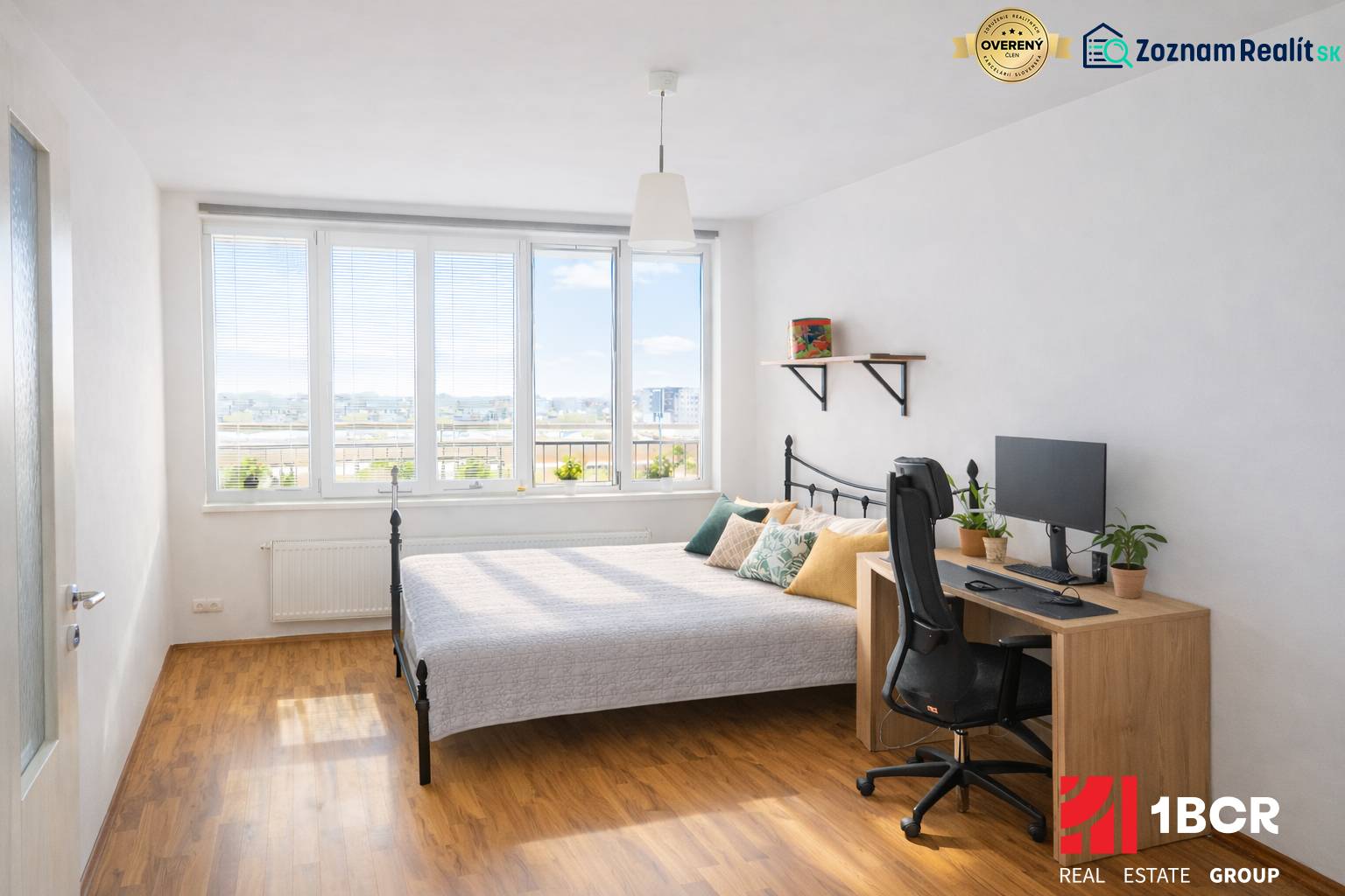 Ložnice v dvoupokojovém bytě s postelí, stolem, počítačem a podlahou s dřevěným dekorem.