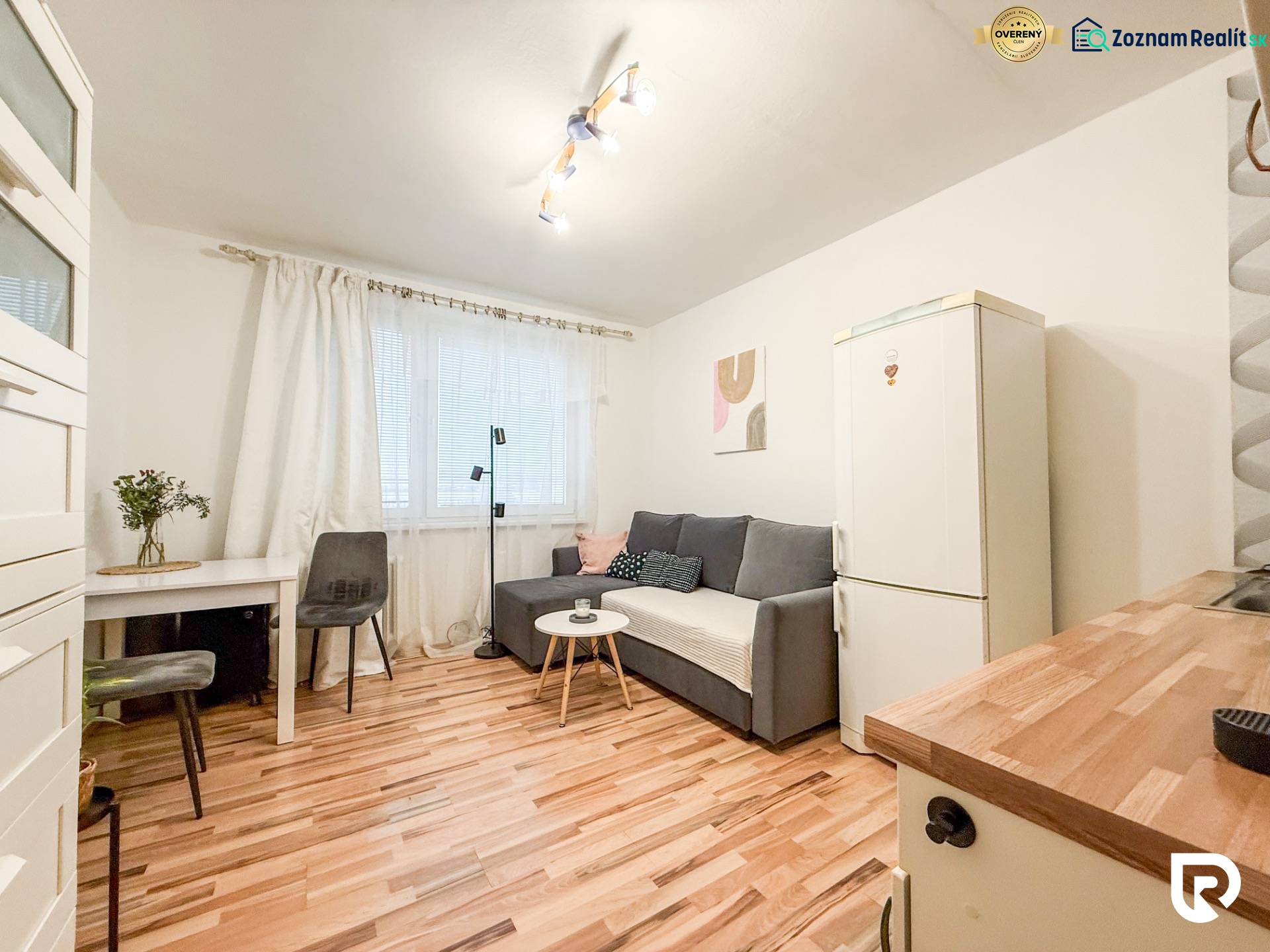 Garsonka s podlahou s dřevěným dekorem, šedou sedačkou a bílou lednicí.