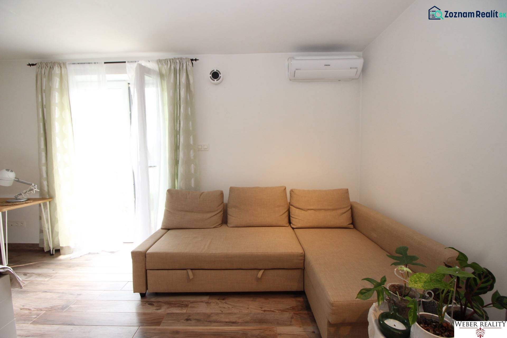 Světlo z okna v 1-pokojovém bytě s pohovkou, pracovním stolem a podlahou s dřevěným dekorem.