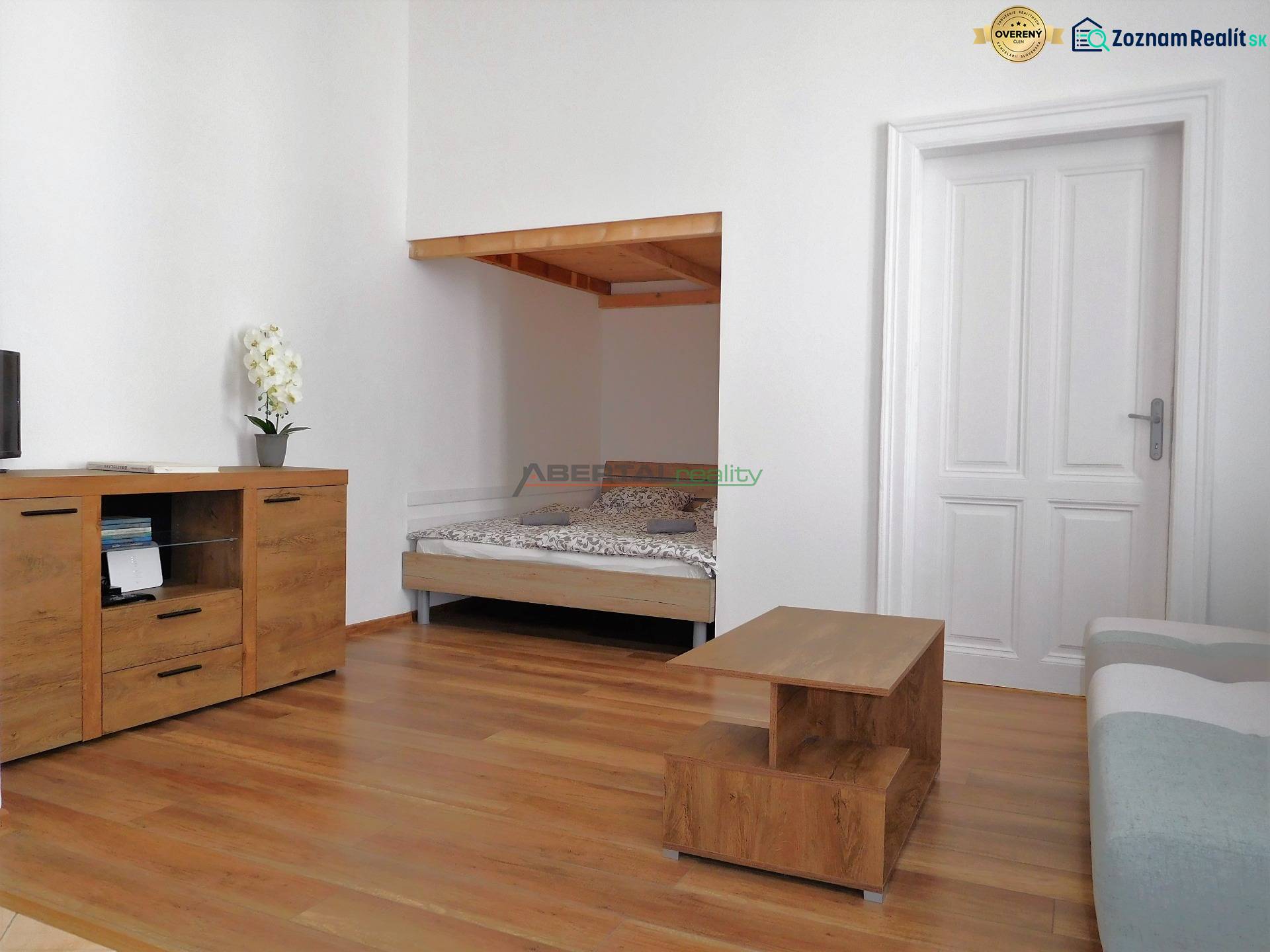 Obývací pokoj dvoupokojového bytu s postelí, skříňkou a podlahou s dřevěným dekorem.