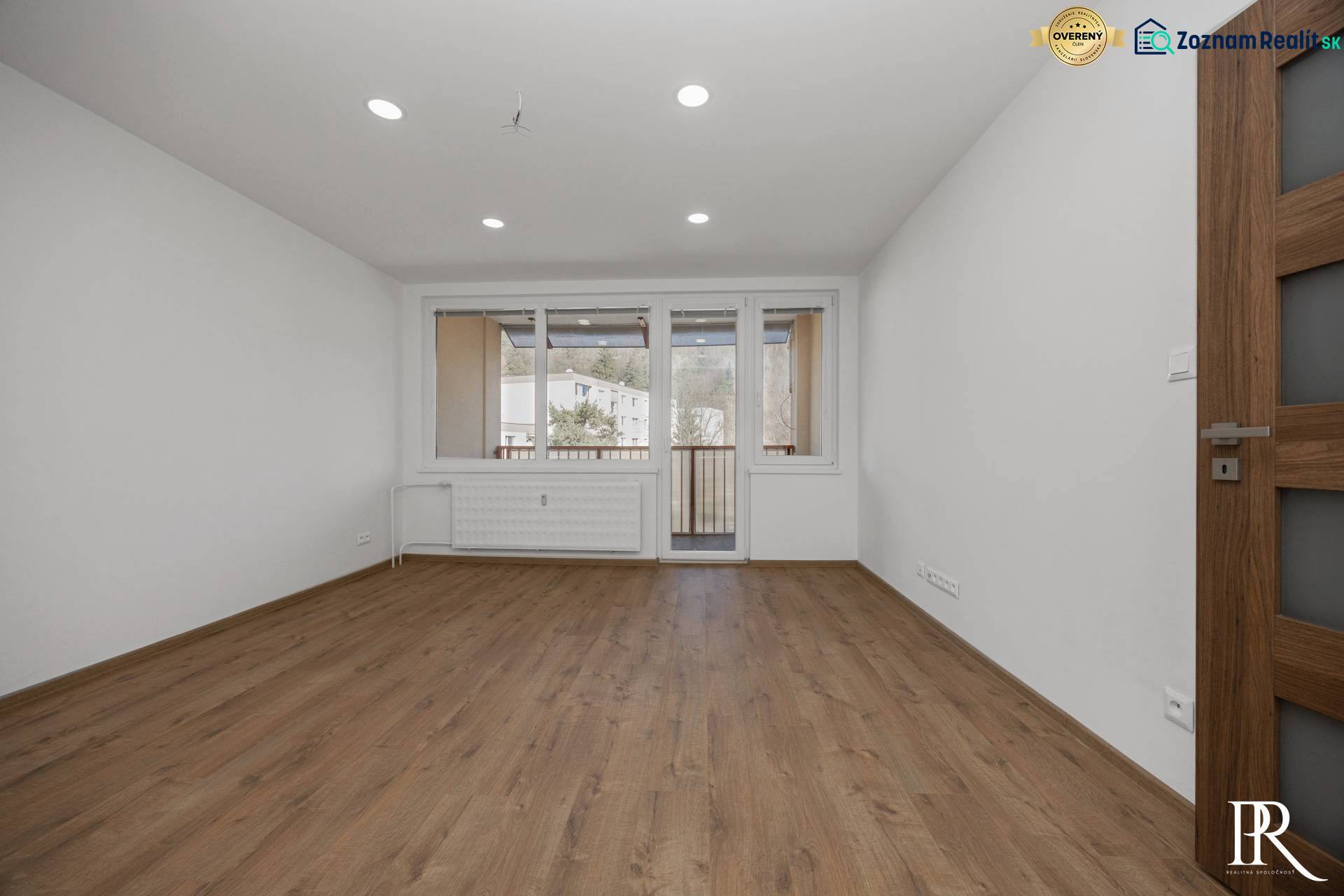 Světlý pokoj ve 3-pokojovém bytě s podlahou s dřevěným dekorem a balkonovými dveřmi.