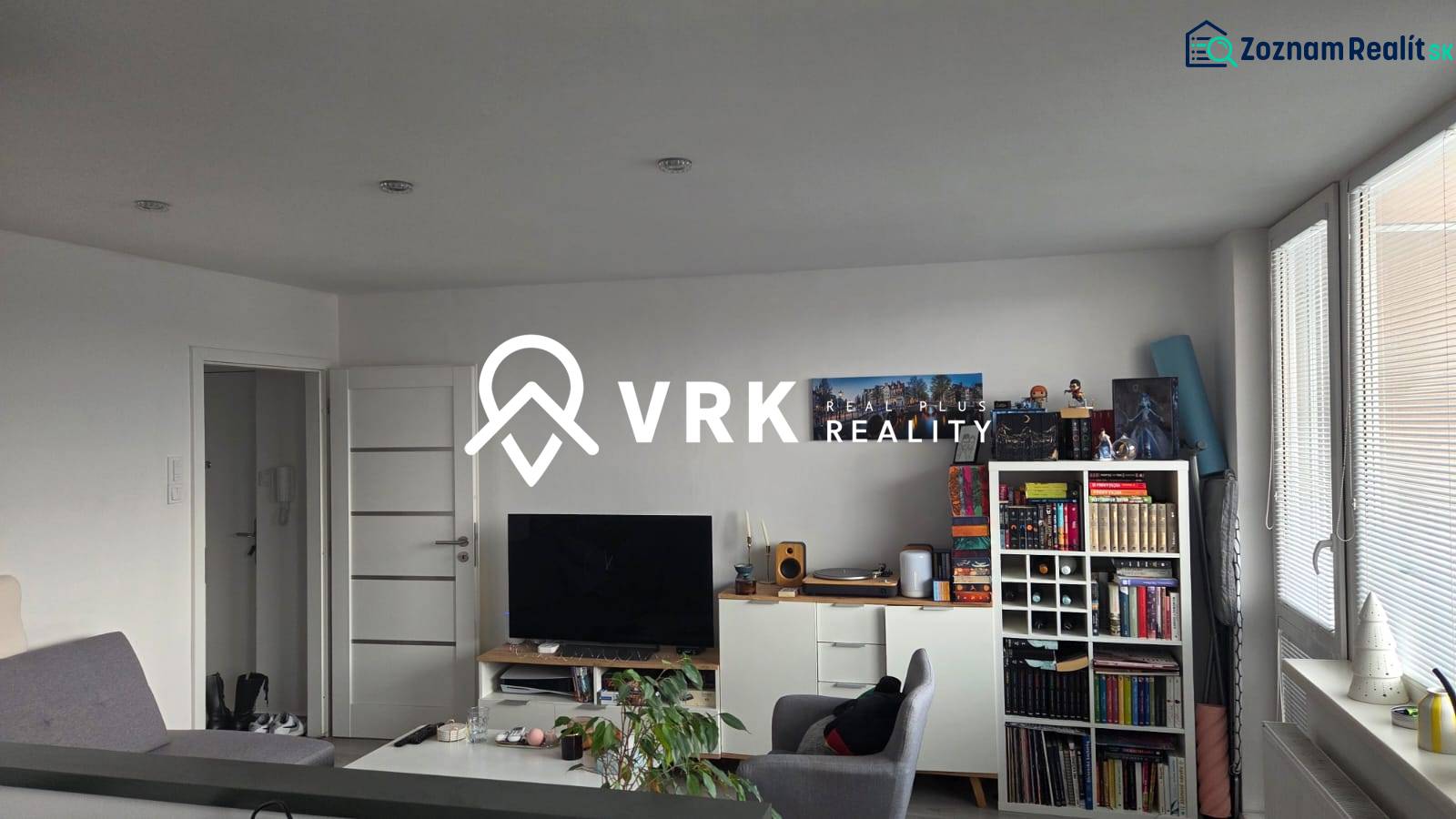 Obývací pokoj třípokojového bytu s knihovničkou, televizorem a světlým nábytkem.