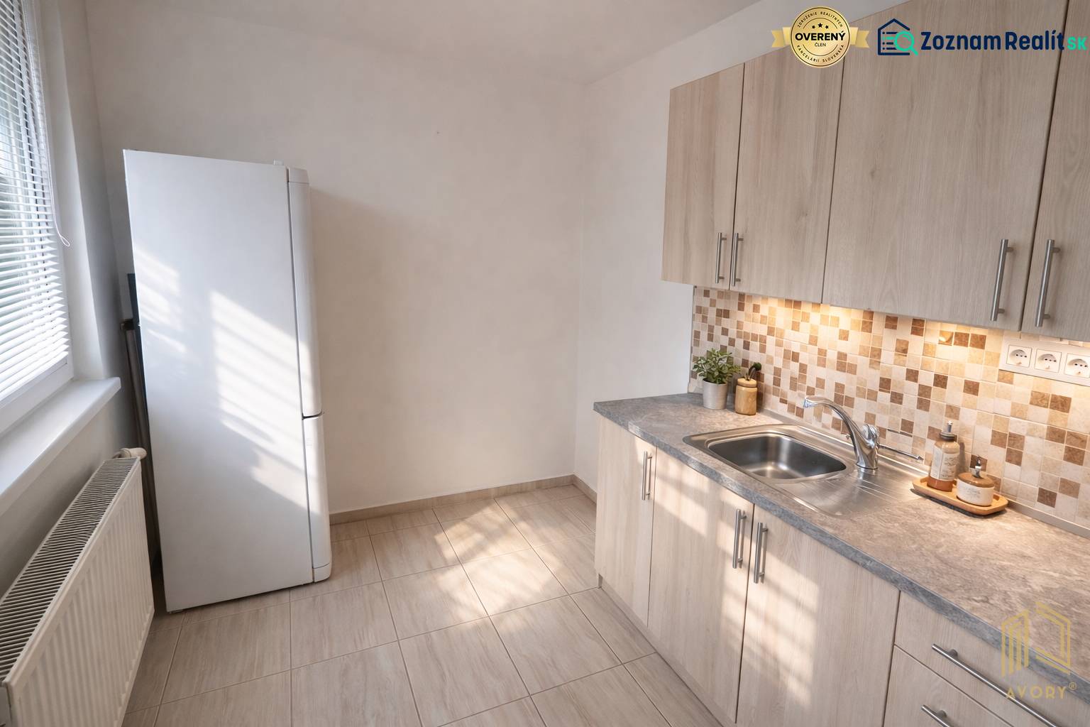 Kuchyně v 2-pokojovém bytě s dlažbou a světlým nábytkem, včetně ledničky.