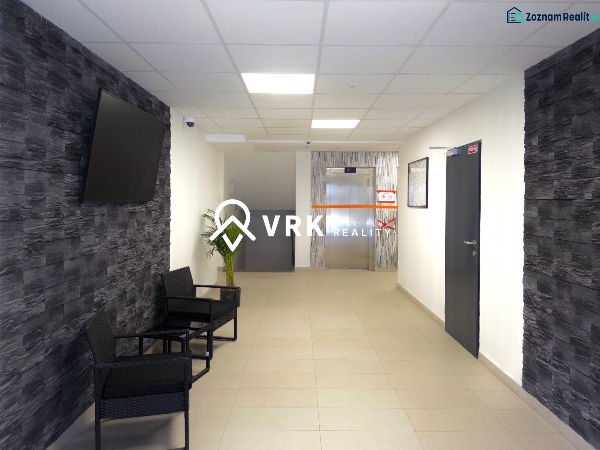 Předsíň v 3-pokojovém bytě s TV na stěně a výtahem v pozadí.