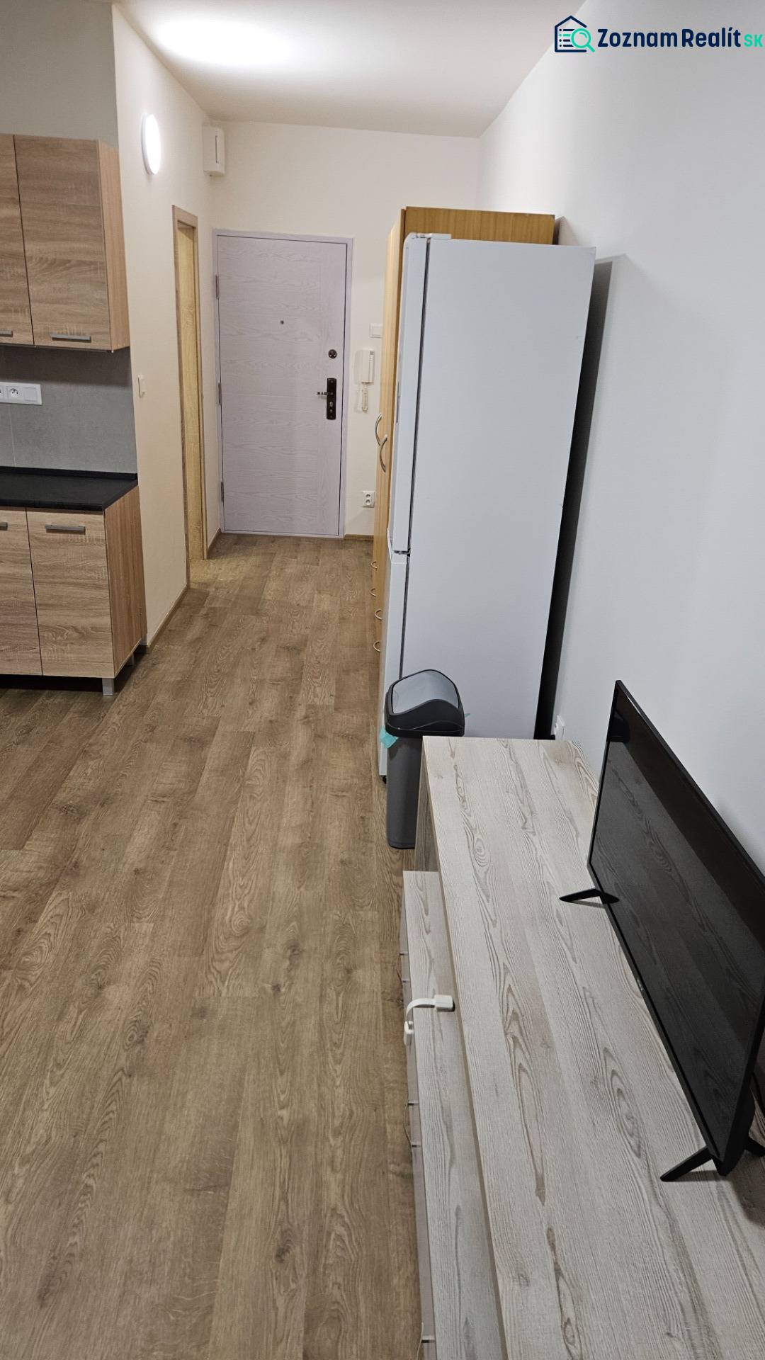 Garsonka s podlahou s dřevěným dekorem, kuchyňskou linkou, lednicí a televizí.