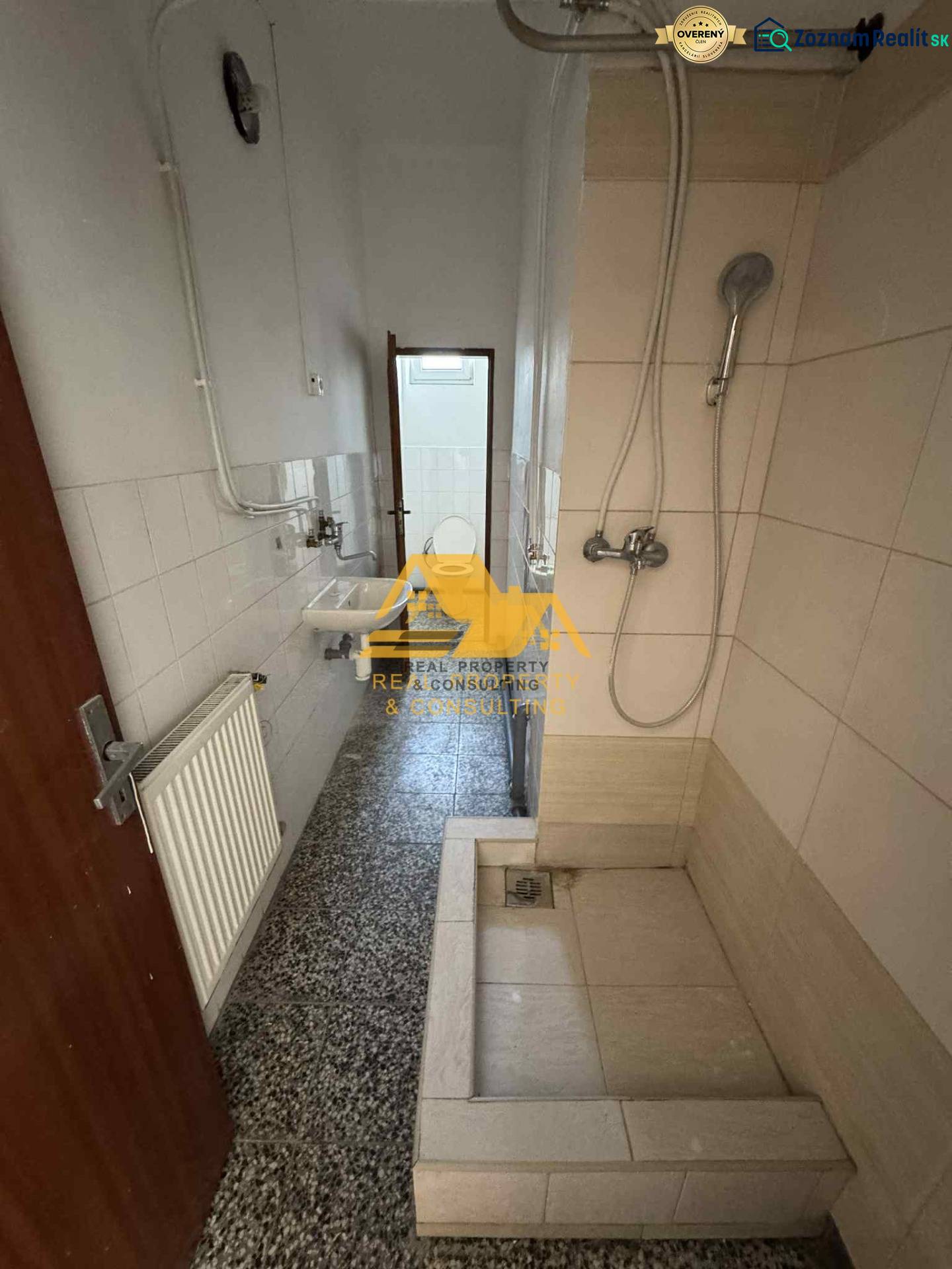 Koupelna v garsonce s jednoduchým sprchovým koutem, umyvadlem a kachličkami.