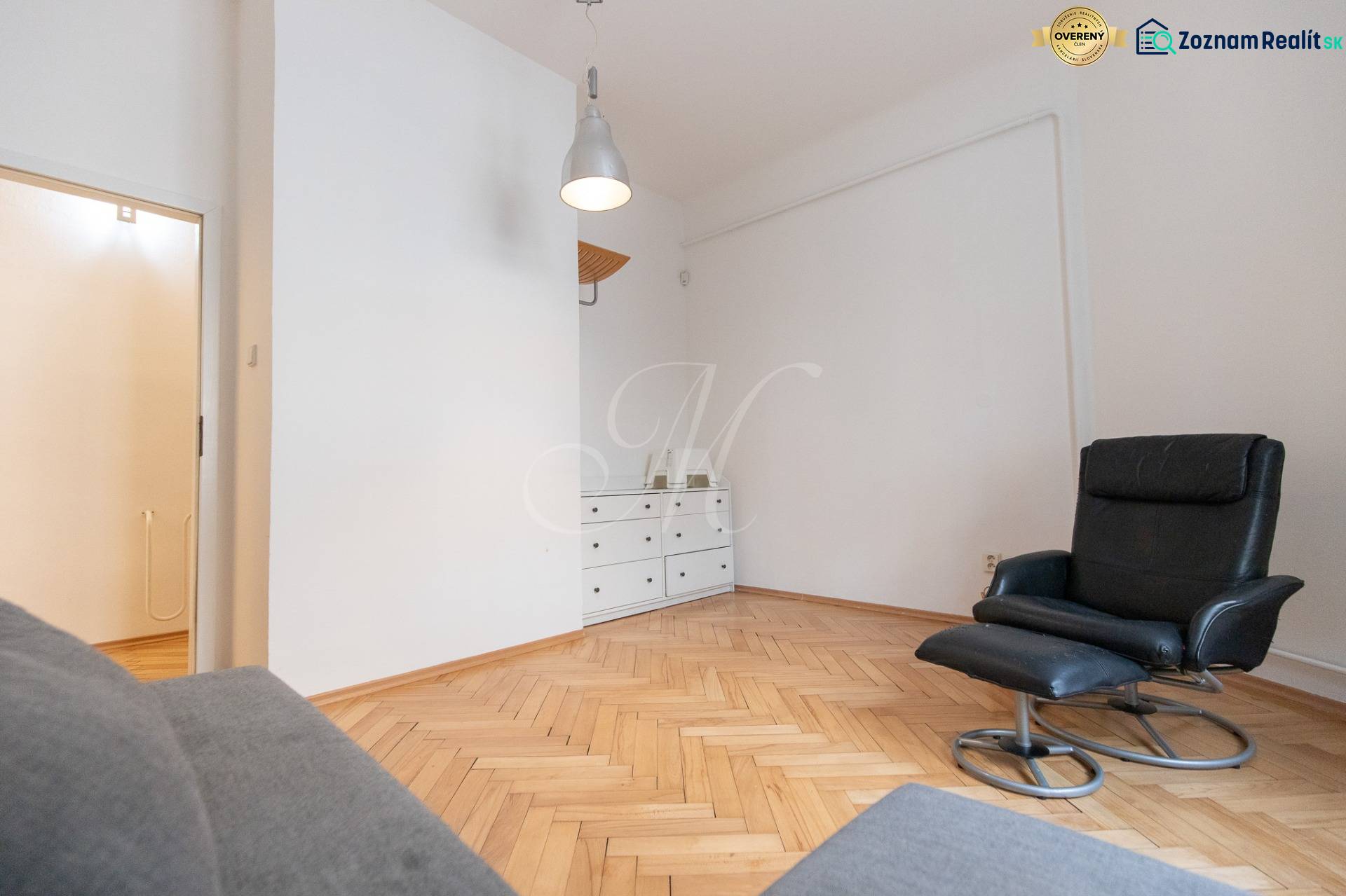 Interiér 4-pokojového bytu s křeslem, bílou komodou a podlahou s dřevěným dekorem.