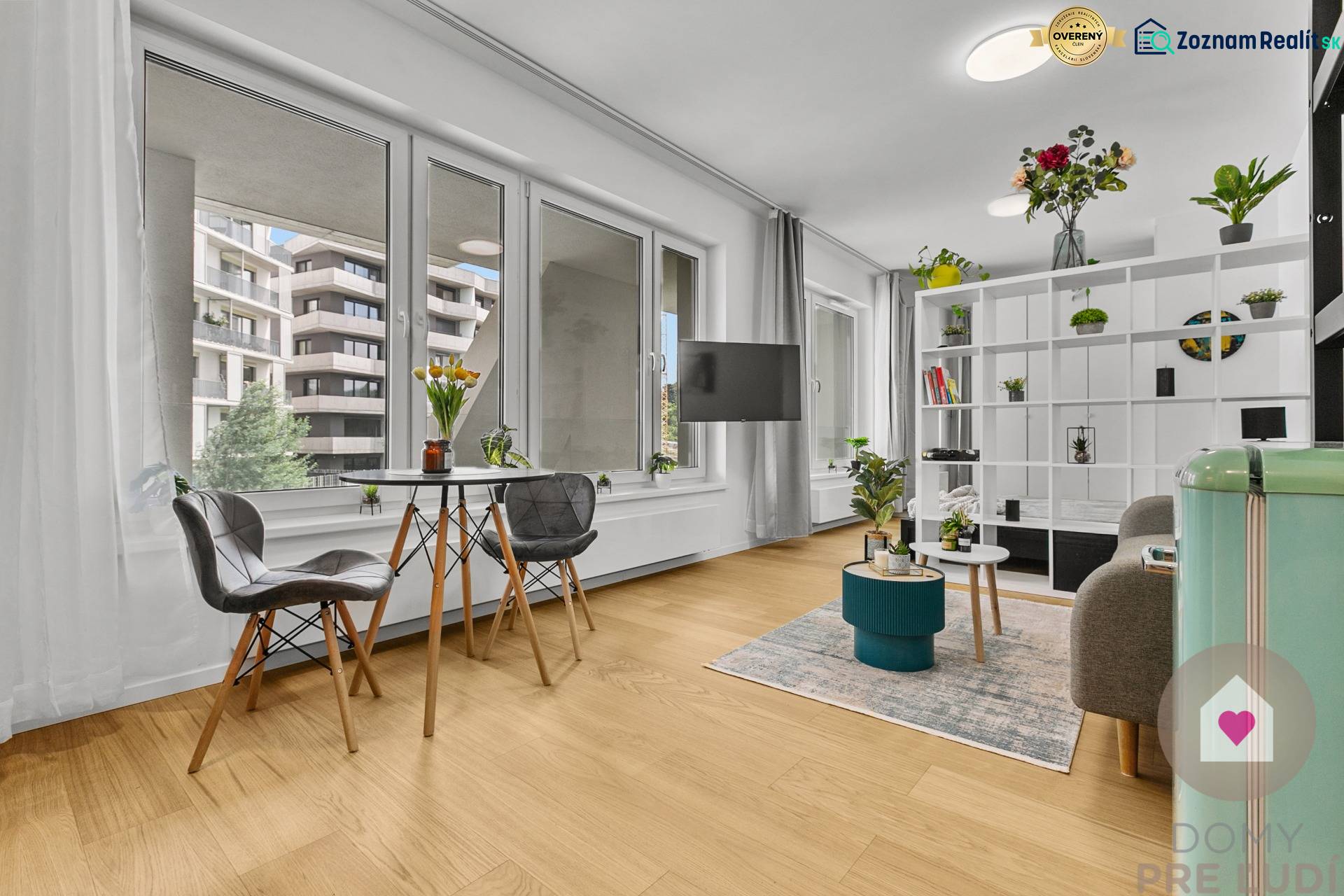 Obývací pokoj jednoizbového bytu s velkými okny, podlahou s dřevěným dekorem a rostlinami.