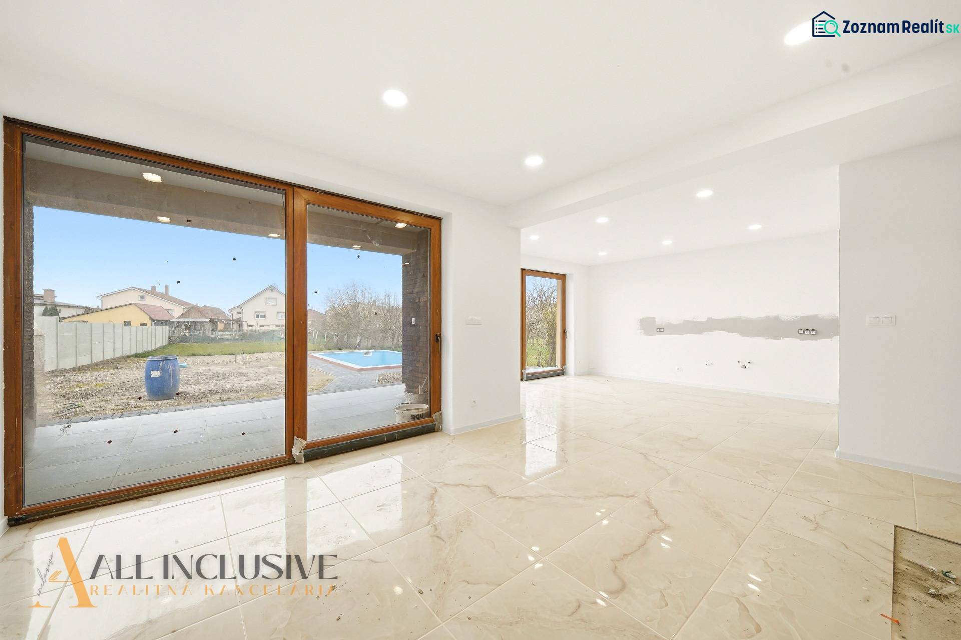 Prostorný interiér rodinného domu s velkými okny a výhledem na zahradu s bazénem.