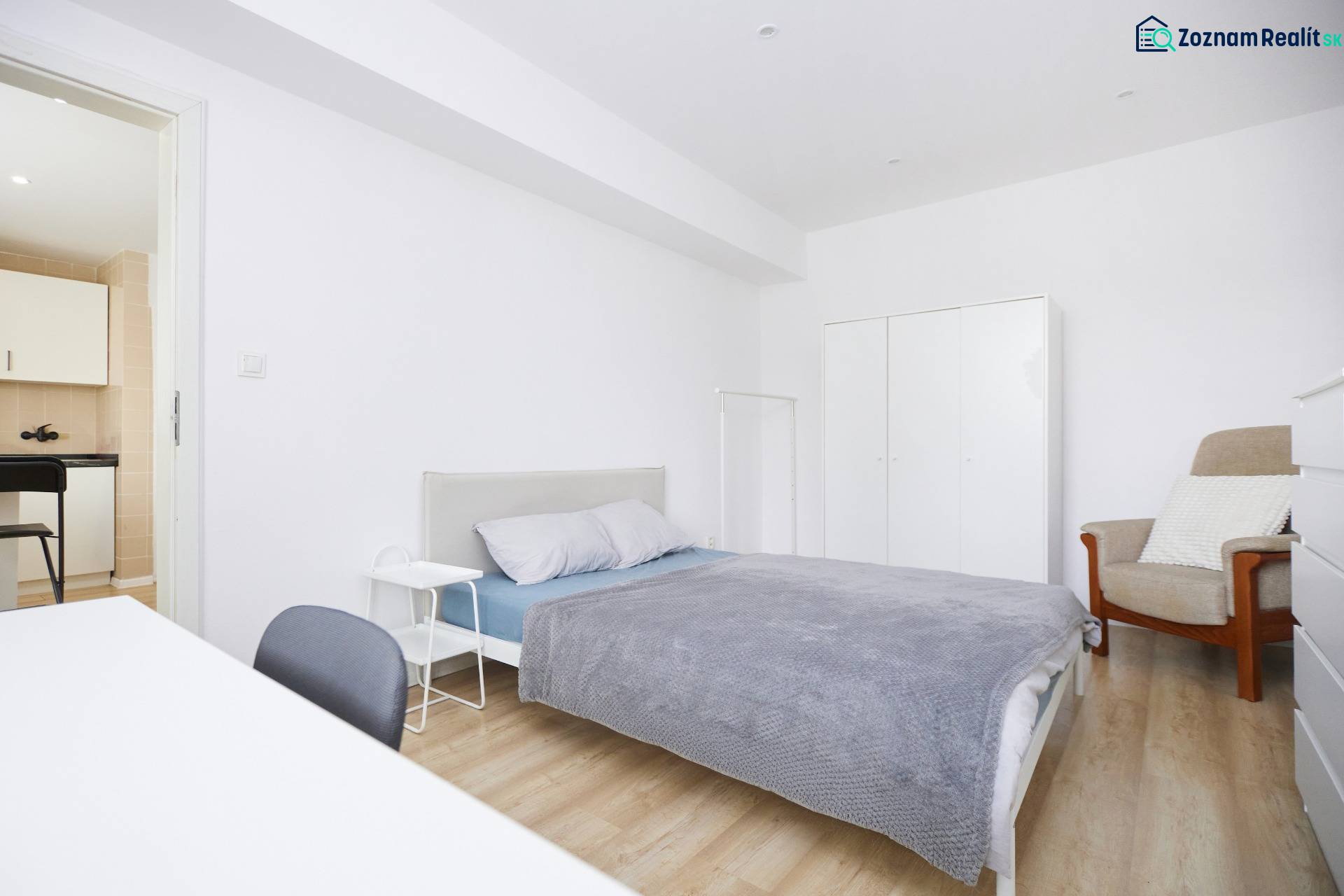 Ložnice dvoupokojového bytu s postelí, skříní a podlahou s dřevěným dekorem.