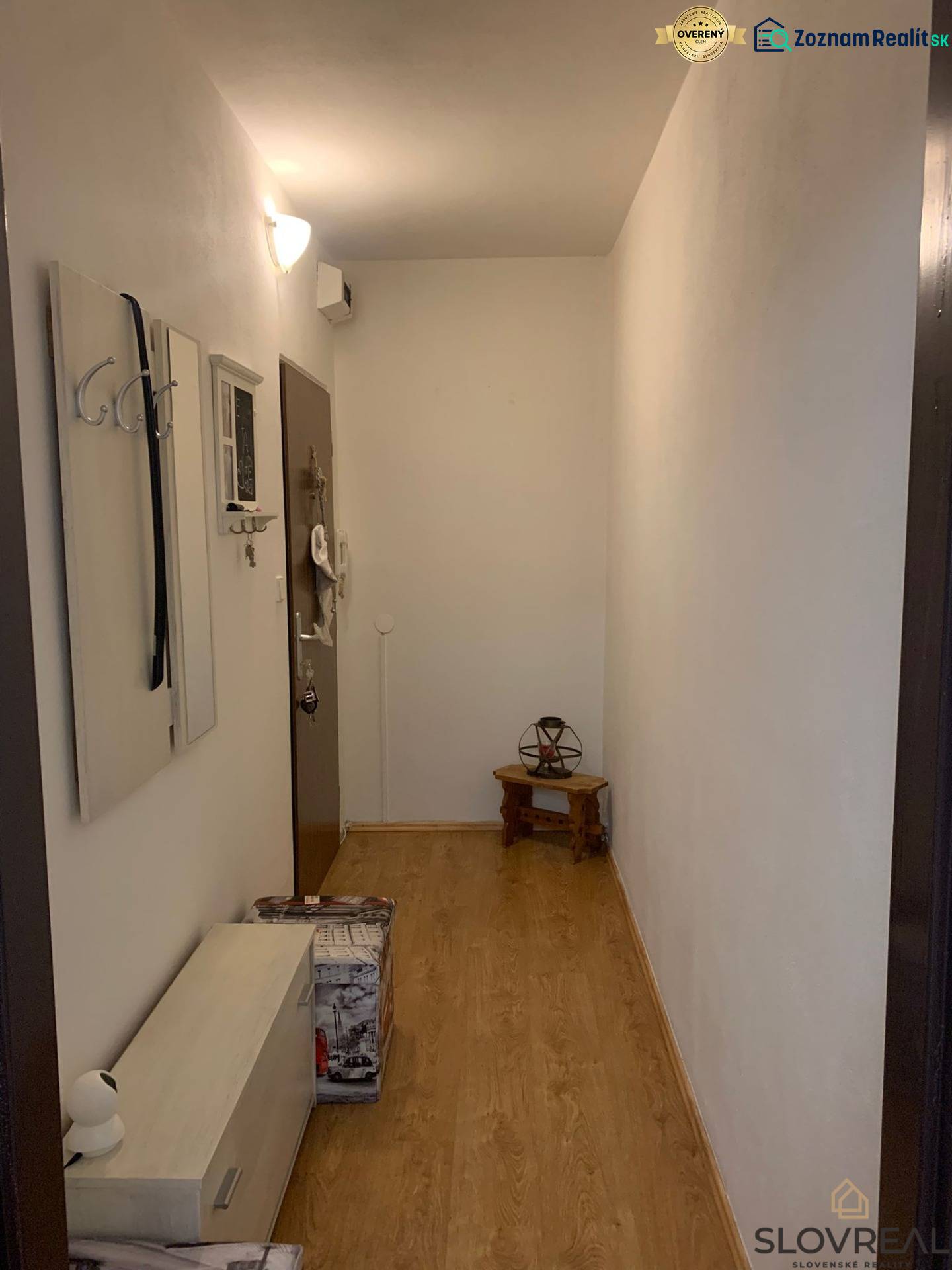 Úzká předsíň s bílými stěnami, podlaha s dřevěným dekorem a minimalistickým nábytkem.