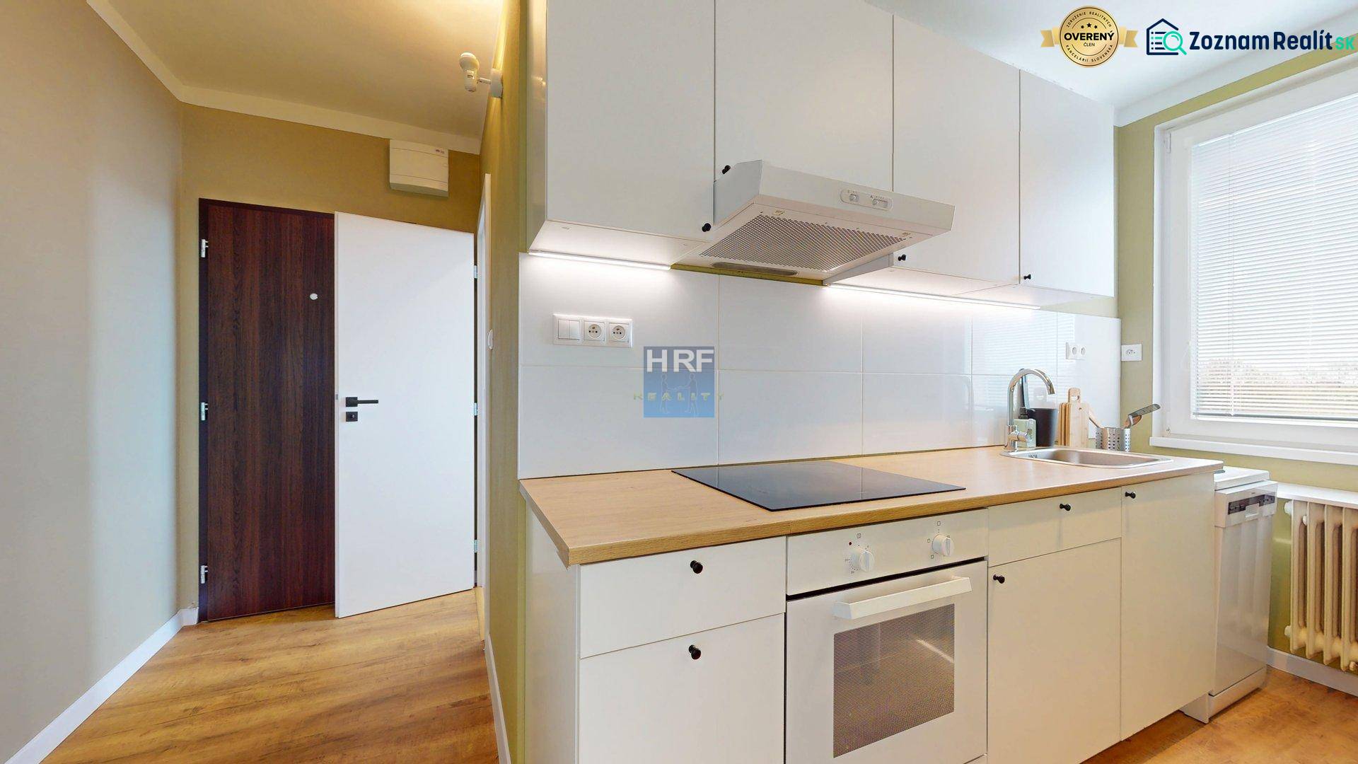 Kuchyně v 1-pokojovém bytě s bílými skříňkami, podlaha s dřevěným dekorem, lednice, varná deska.