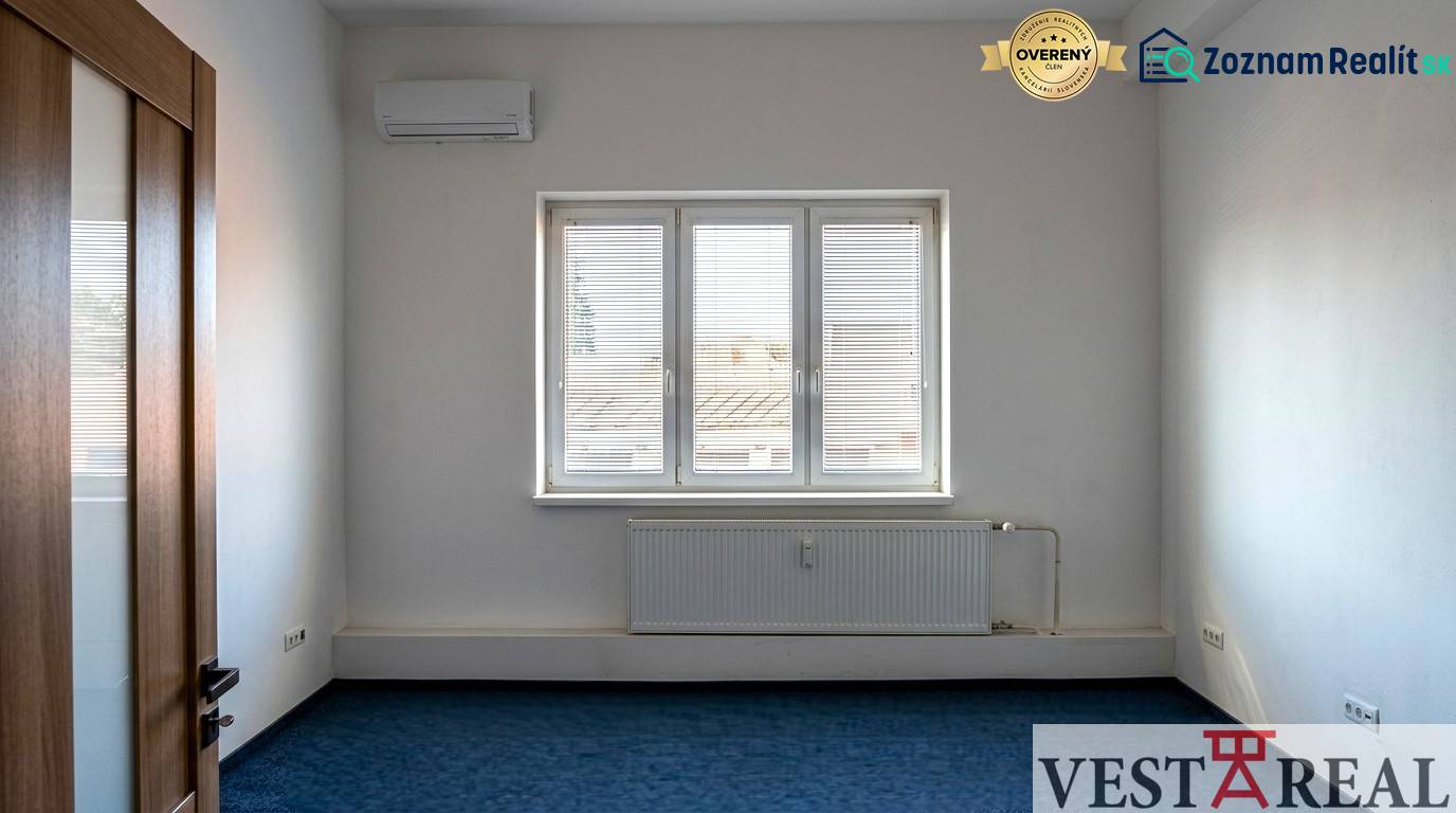 Místnost s klimatizací a oknem v Kancelářích, modrý koberec, bílé stěny.