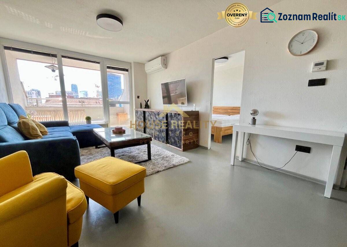 Obývací pokoj třípokojového bytu s pohovkou, televizí a balkonem.