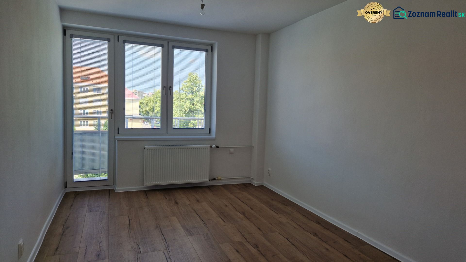 Pokoj ve 3pokojovém bytě s podlahou v dřevěném dekoru, velkým oknem a radiátorem.