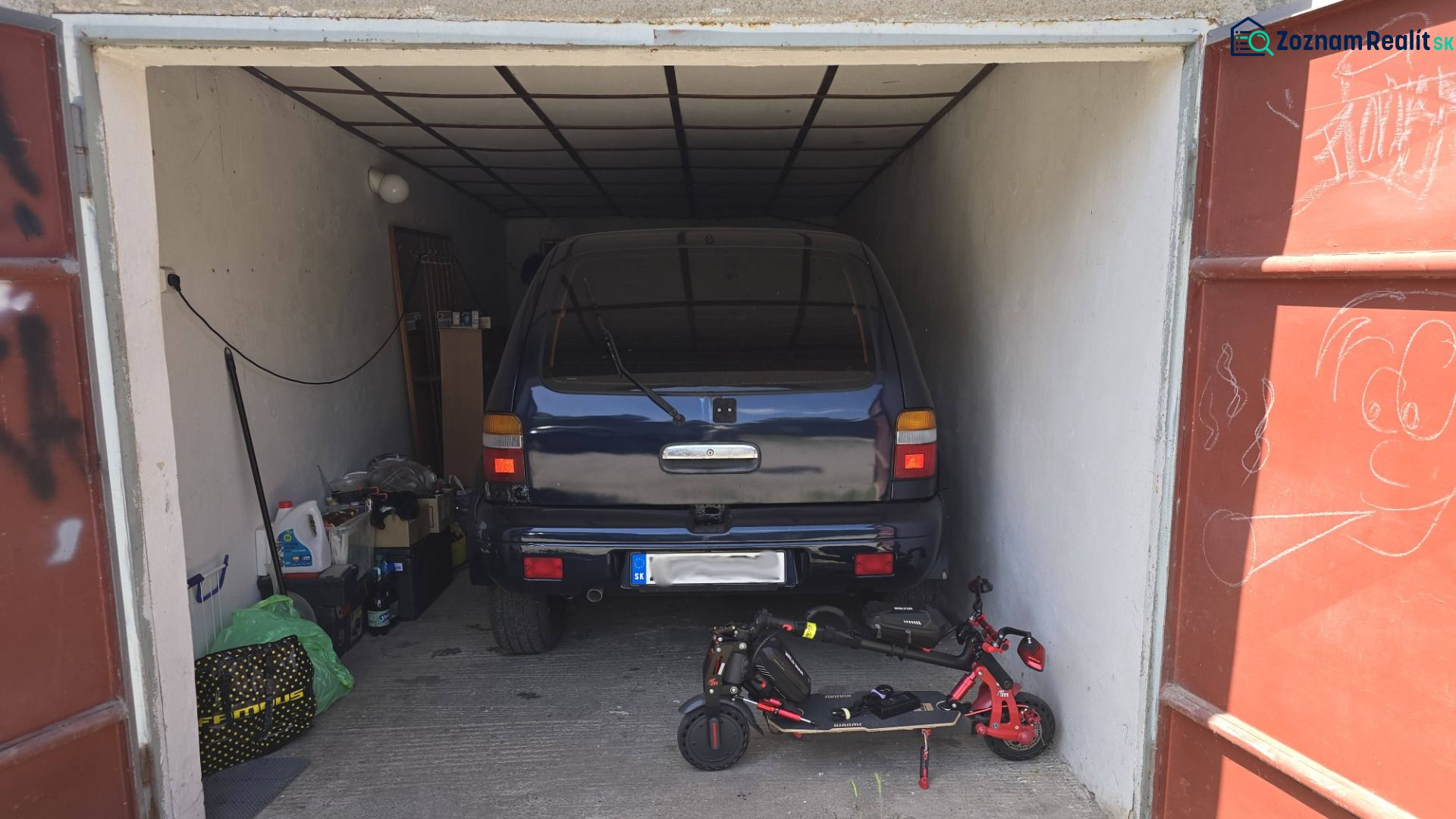 Garáž s autem a elektrickou koloběžkou, uloženými předměty a kresbou na dveřích.