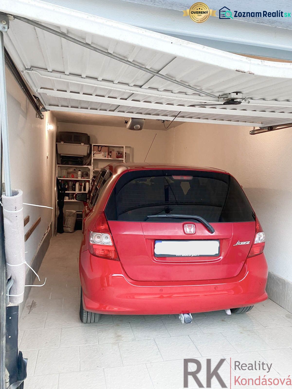 Červené auto zaparkované v garáži s regály na stěně, podlaha z dlažby.